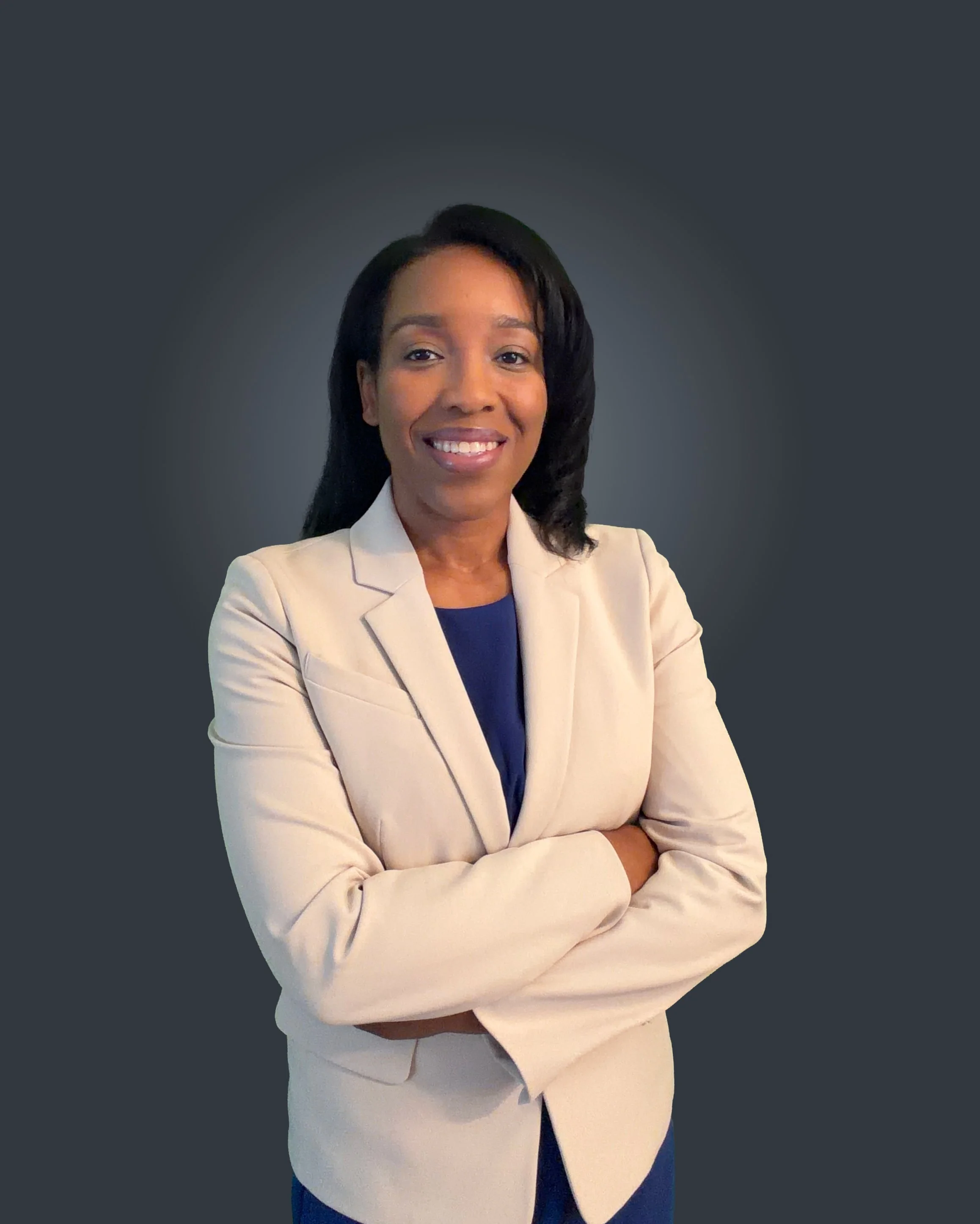 A woman with medium brown skin and black hair, smiling, dressed in a beige blazer and blue top, with arms crossed, standing against a dark gray background.