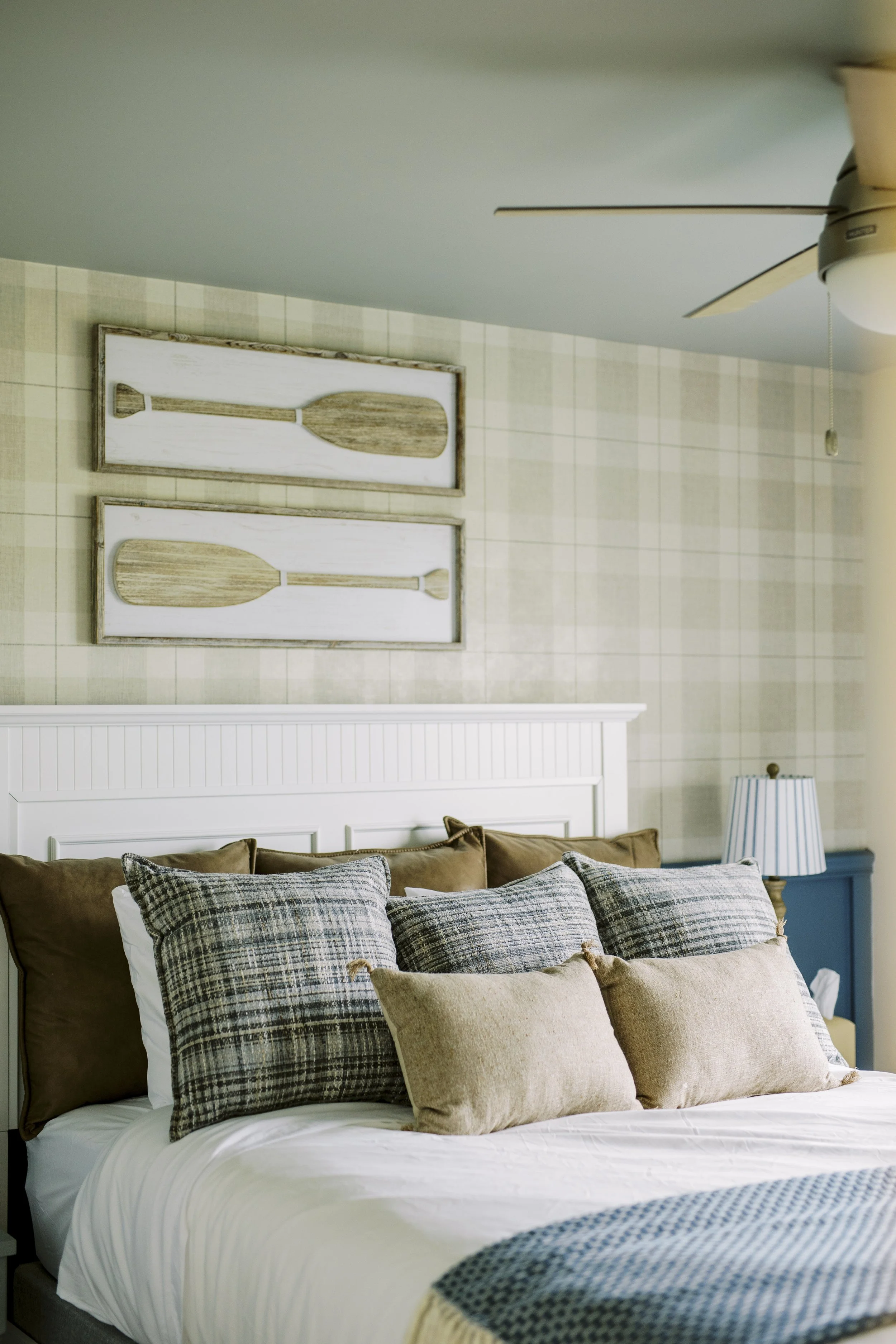 A cozy bedroom with a white bed covered in plaid and neutral-colored pillows, a blue nightstand with a striped lamp, and wall art featuring wooden oars hanging on a checkered wallpapered wall. There is a ceiling fan with a light in the top right corner.