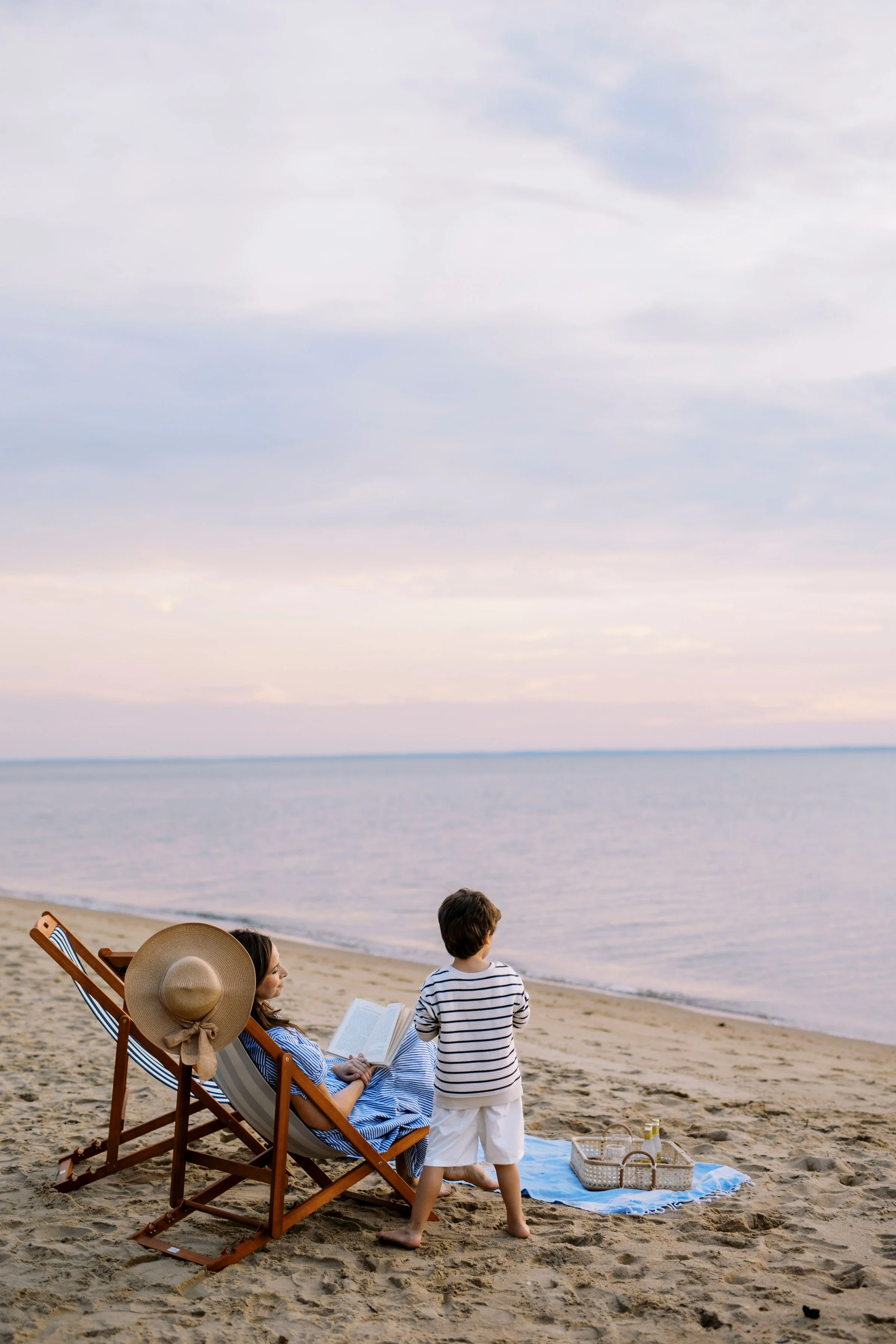 People relaxing on a beach, with one woman sitting in a chair reading a book, a child standing nearby, and another person partially visible. The beach has sand, a calm sea, and a cloudy sky.