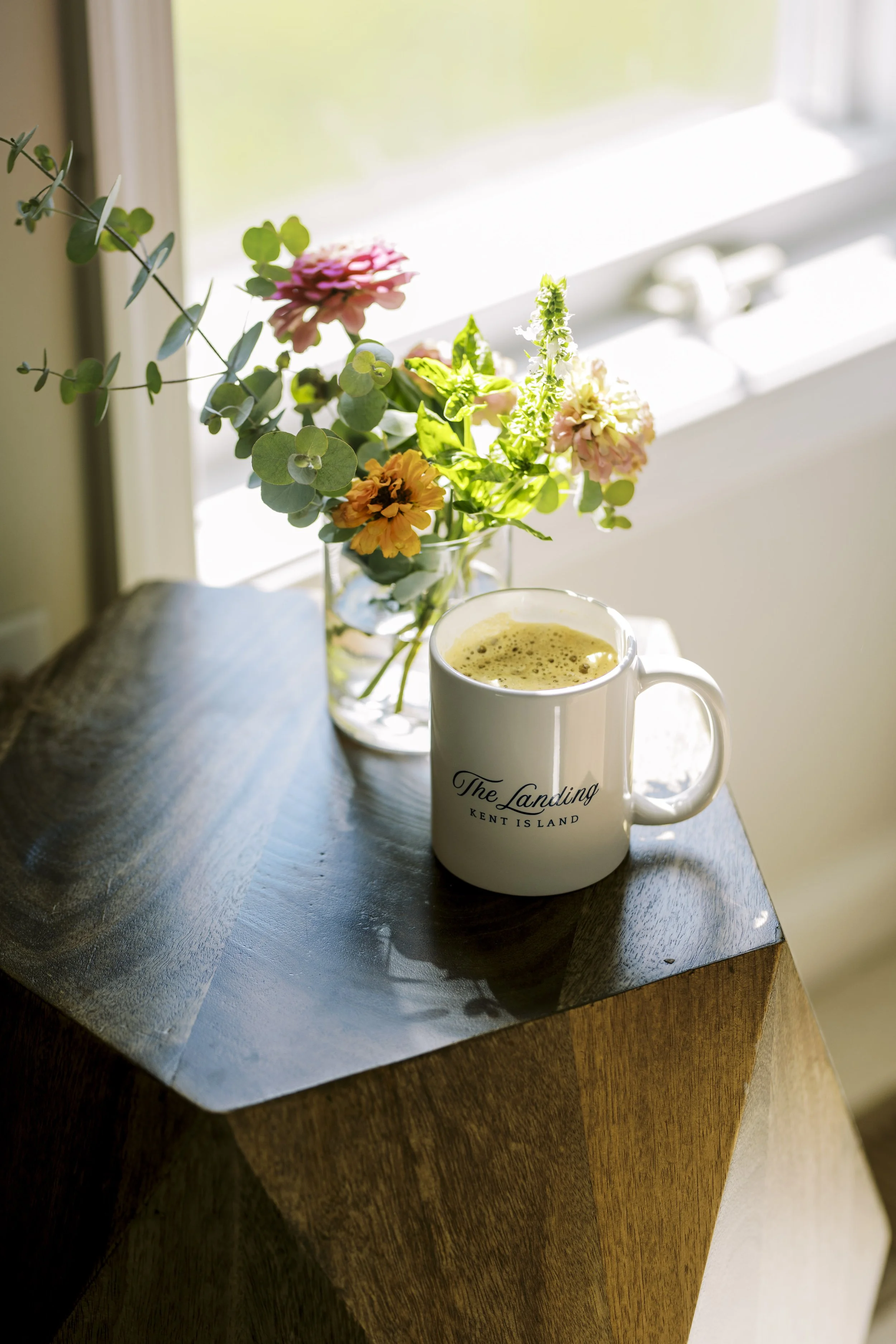 A white mug with the text 'The Landing Kent Island' on a wooden table, next to a glass vase of pink, orange, and white flowers, near a window with sunlight streaming in.