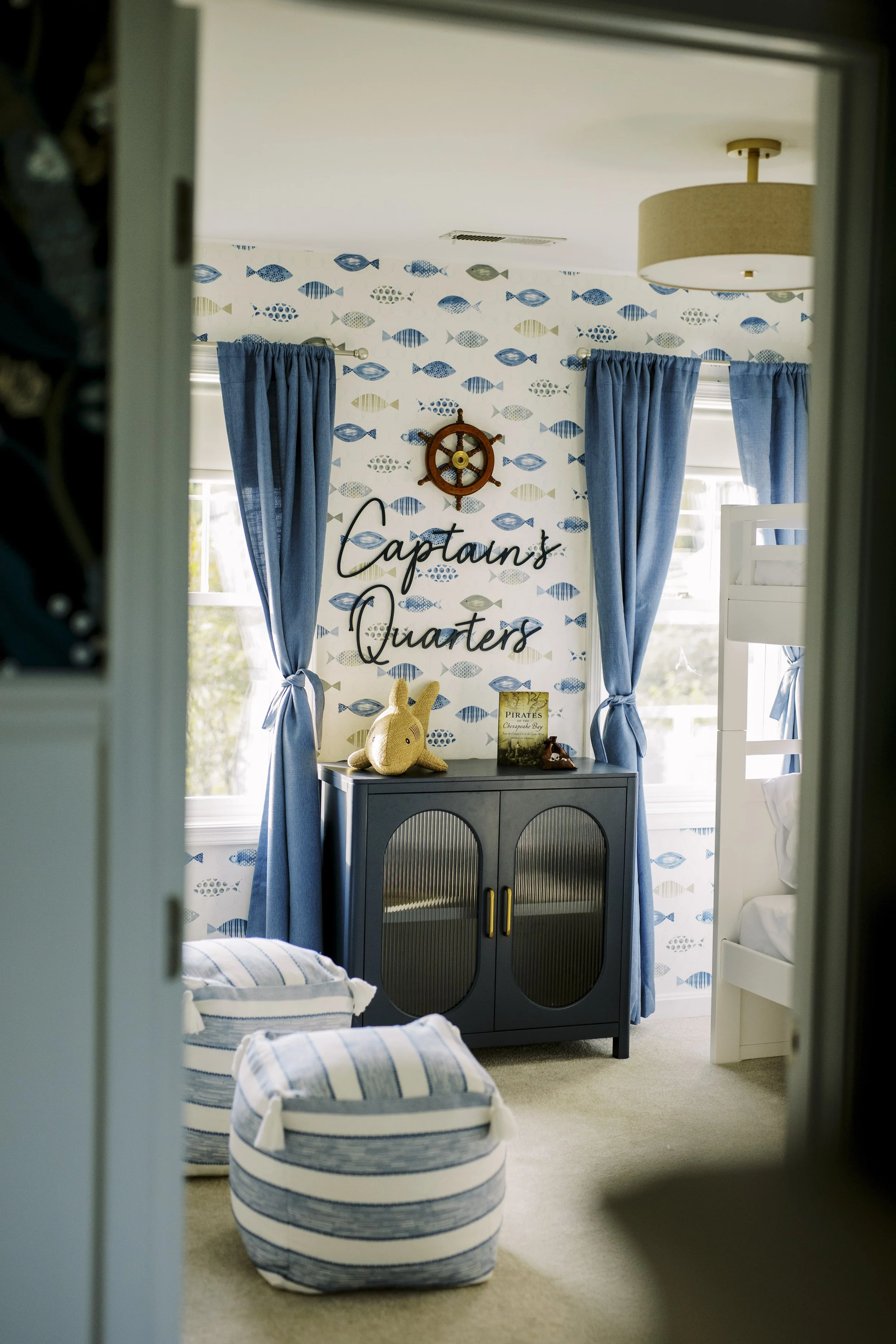 A nautical-themed child's room with fish-patterned wallpaper, blue curtains, and a sign reading 'Captain's Quarters' on a small cabinet. Contains a plush bunny, a book, and striped ottomans.