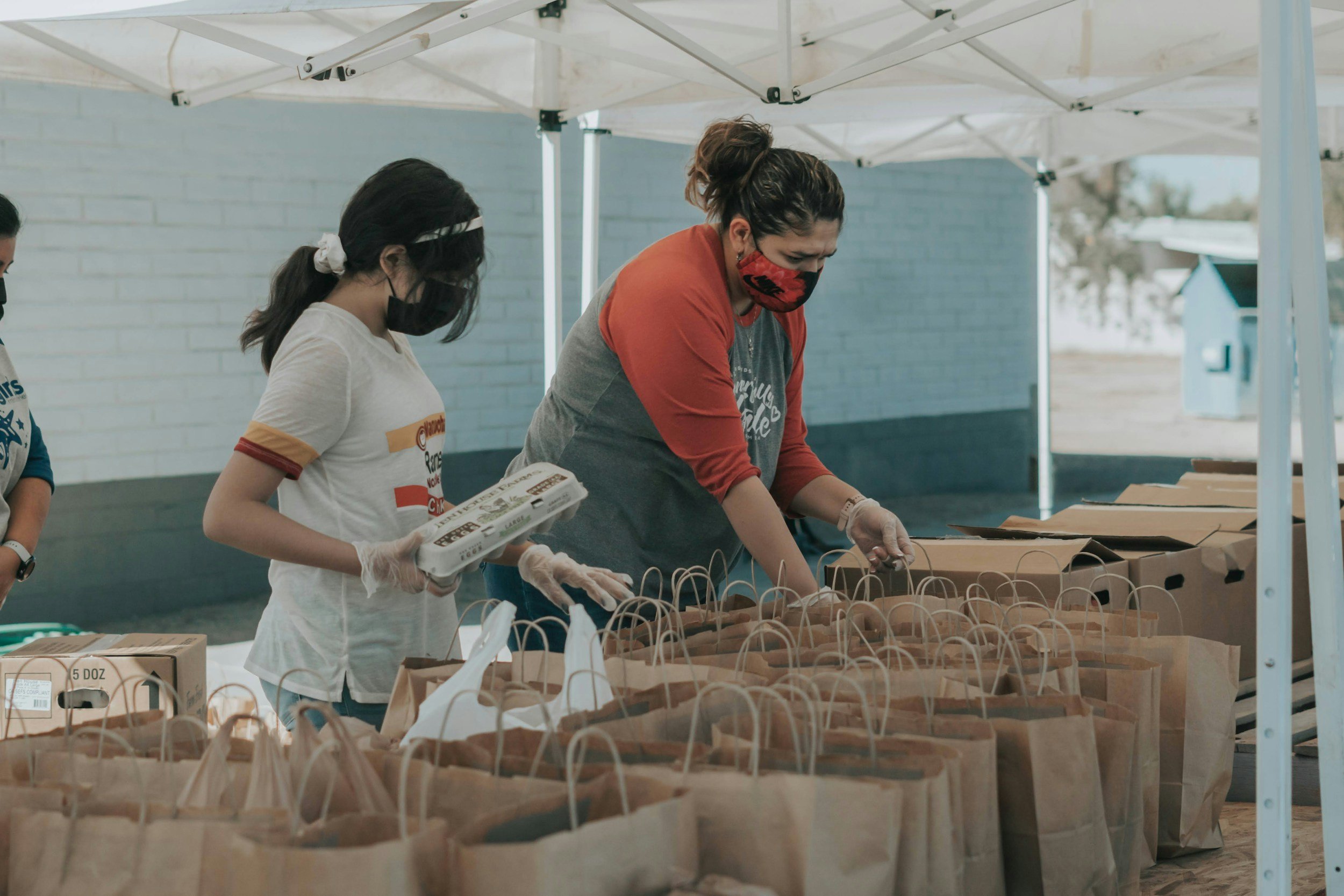 People wearing masks and gloves sorting paper bags under a white canopy at an outdoor event.
