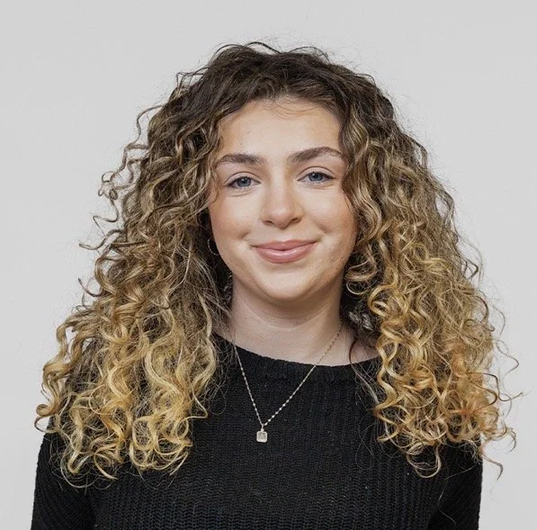 A young woman with long, curly blonde hair, wearing a black top and a necklace with a small pendant, smiling at the camera against a plain light background.