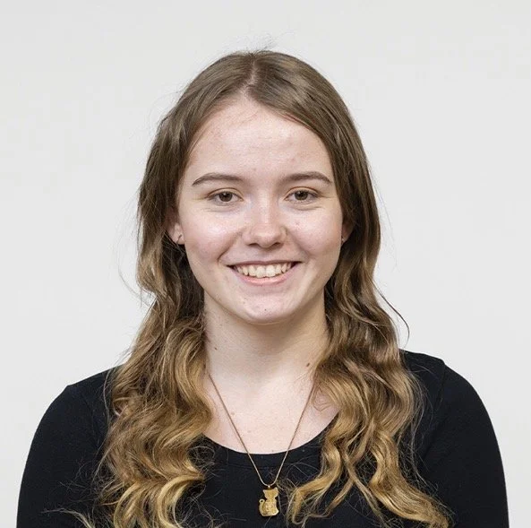 A young woman with long, wavy brown hair smiling at the camera, wearing a black top and a gold necklace with a bear pendant, against a plain white background.