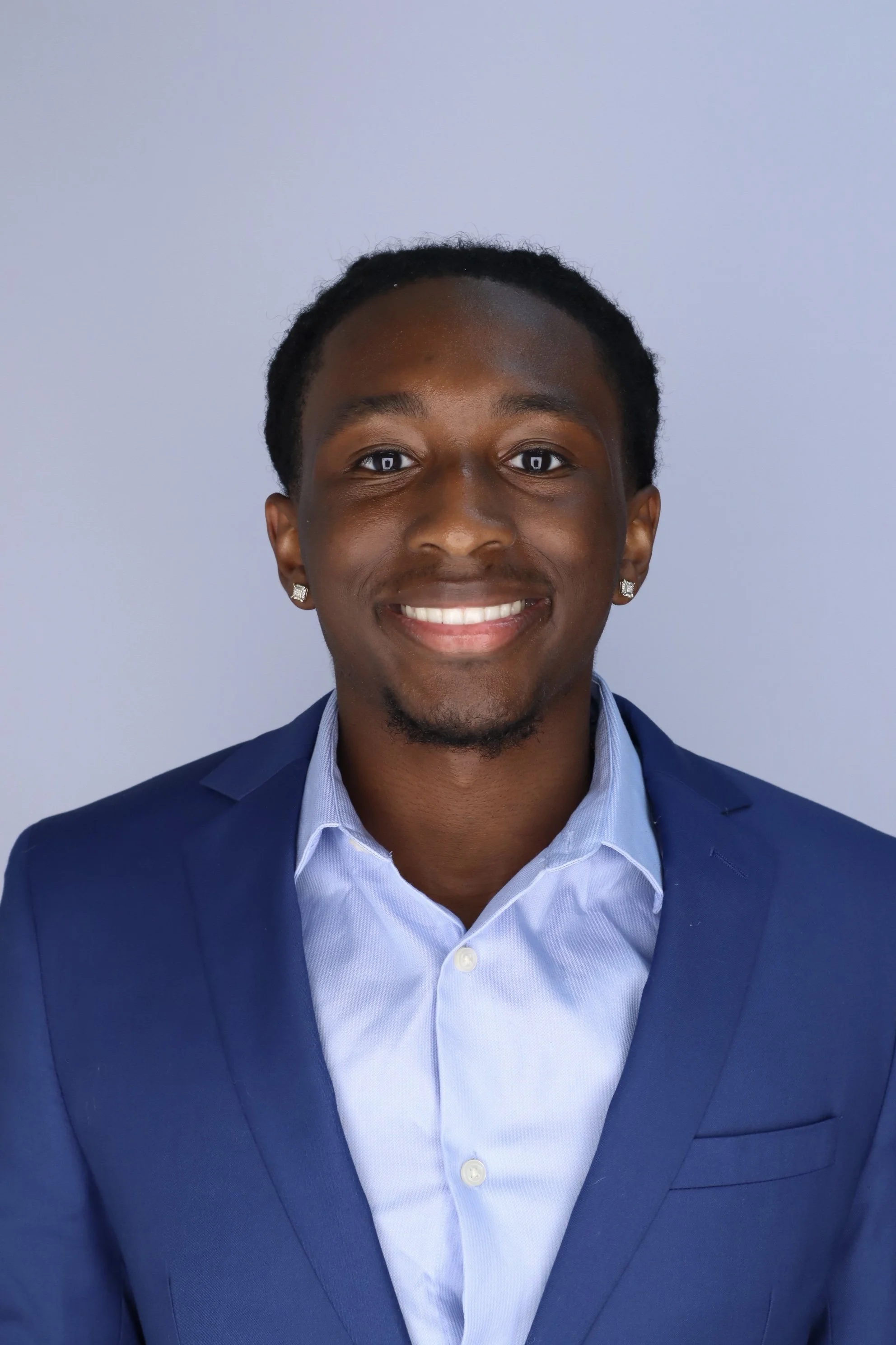 A young man in a blue suit jacket and light blue collared shirt, smiling, with earrings, and short dreadlocks, standing against a plain light gray background.