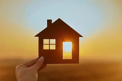 A hand holds a paper cutout of a house in front of a sunset, creating the illusion of the house glowing with sunlight.