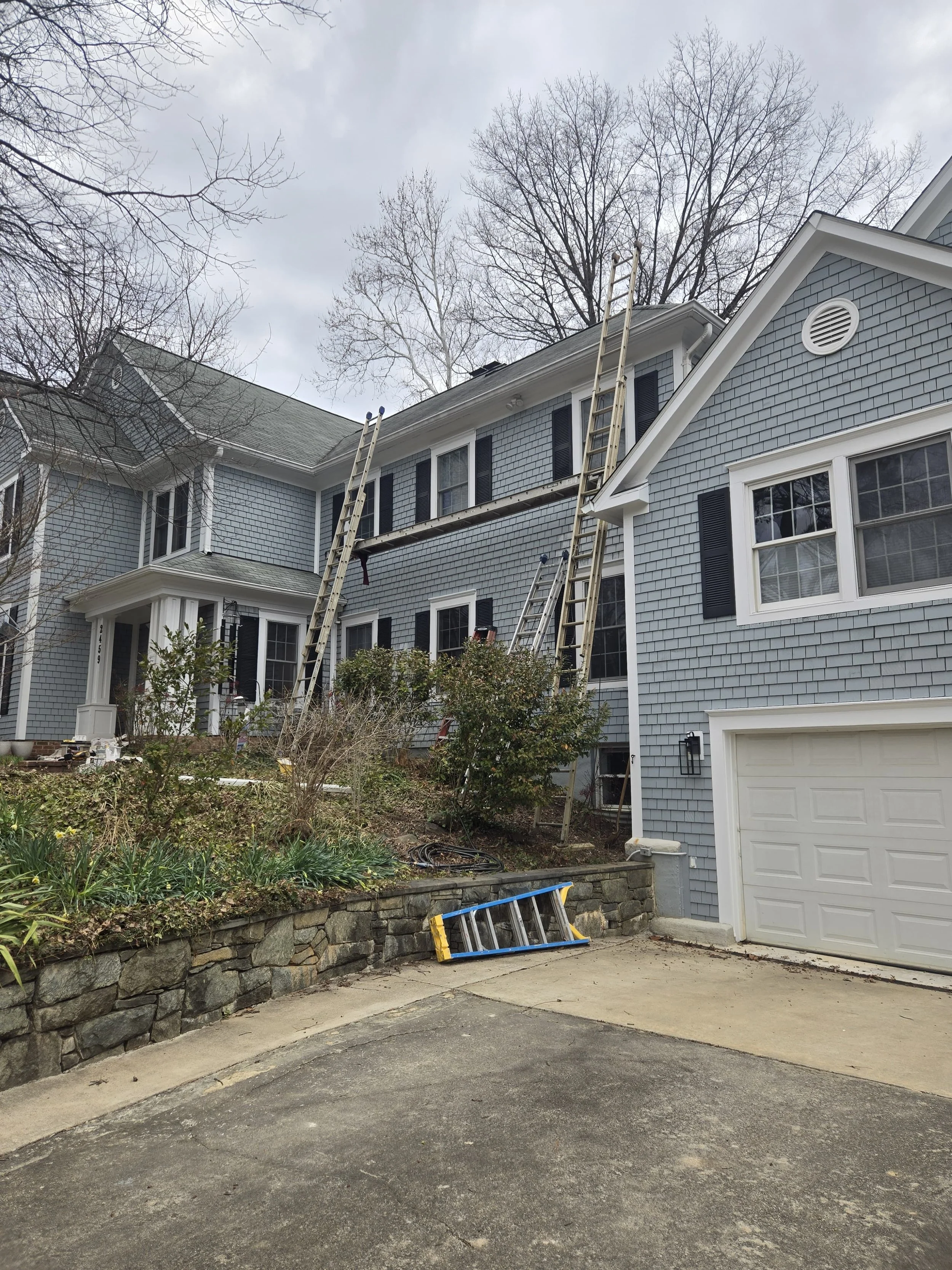 A two-story blue house with black shutters and a white garage door. Ladders leaning against the roof and a tree with no leaves in the background. There is a garden area with plants and a stone retaining wall, and some tools and equipment on the ground.