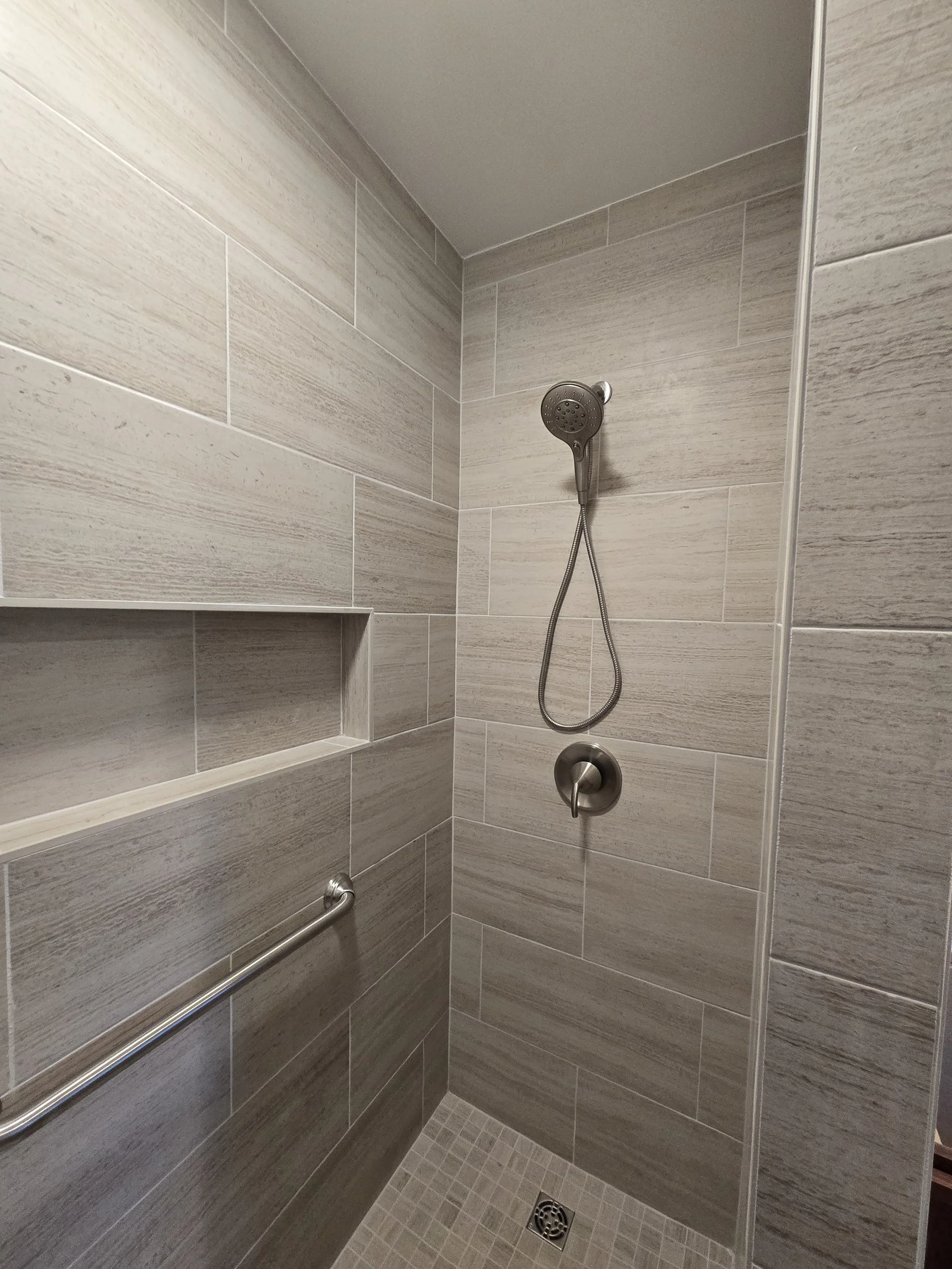 A walk-in shower with gray tiled walls, a handheld showerhead, a grab bar, a niche for toiletries, and a drain on the floor.