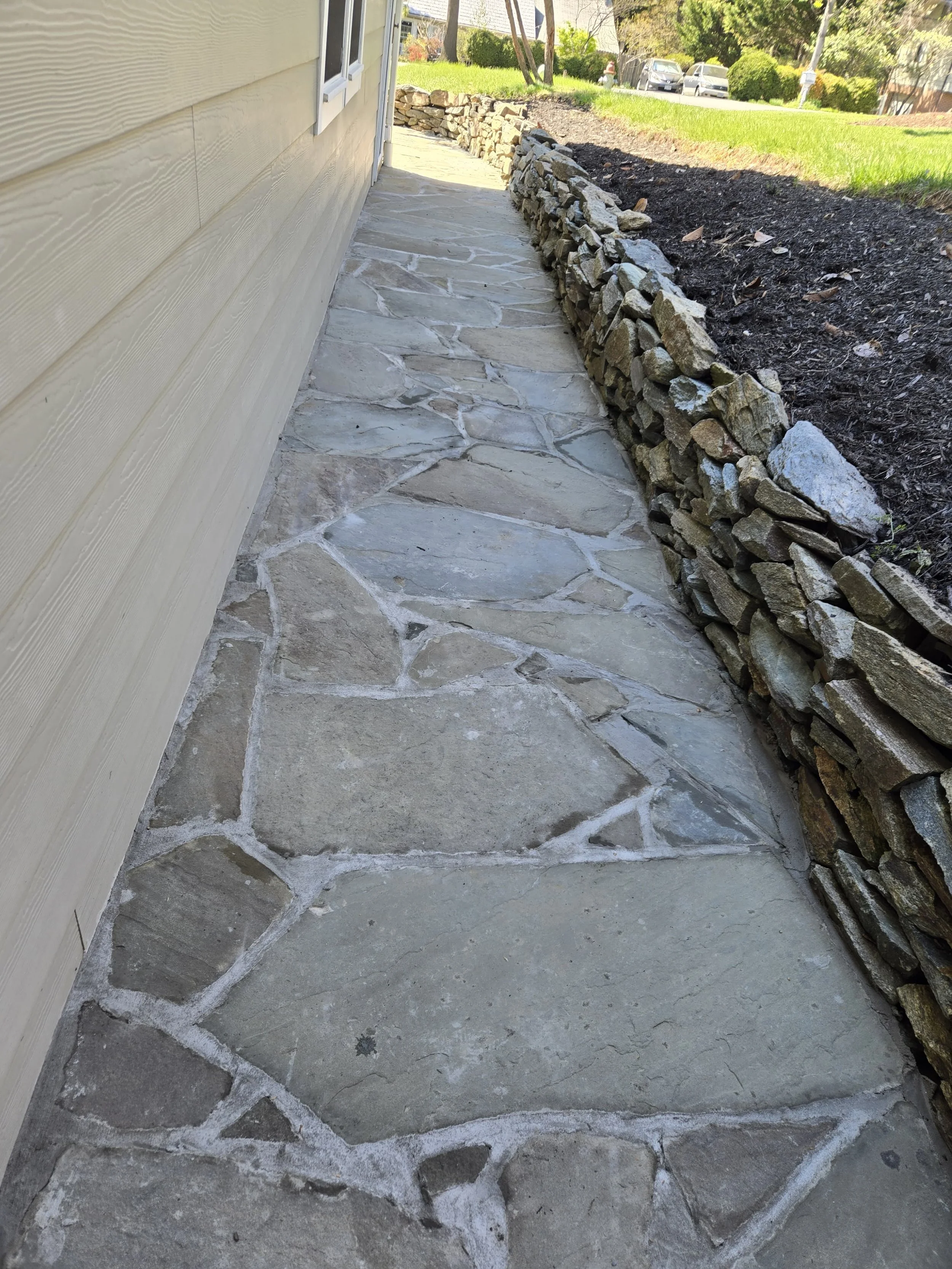 A stone pathway runs alongside a house with beige siding, bordered by a low stacked stone wall on the right. The background shows a grassy hill, trees, and parked cars.