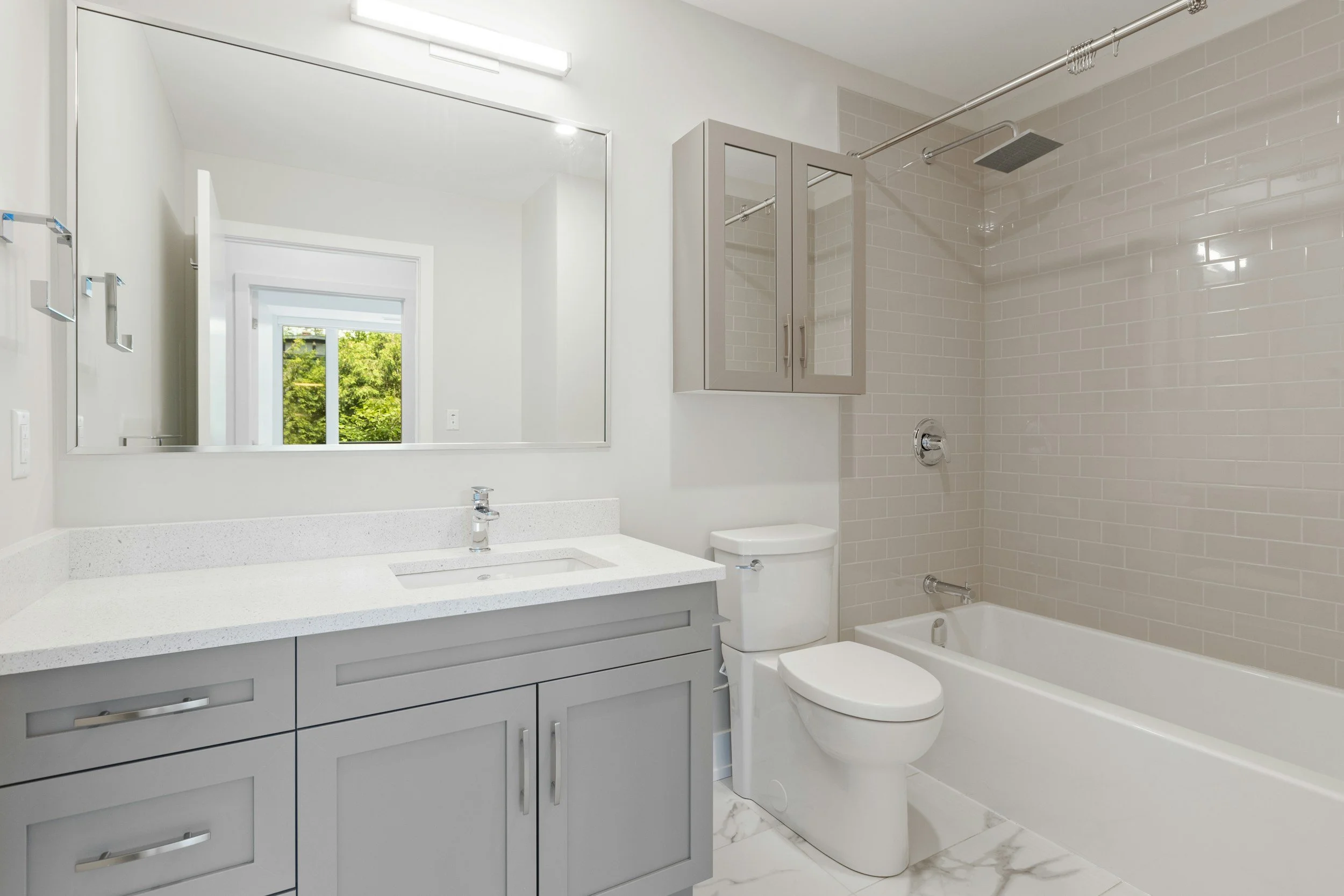 Modern bathroom with a large mirror, a white vanity with grey cabinets, a white toilet, and a walk-in shower with beige subway tiles and a rainfall showerhead.