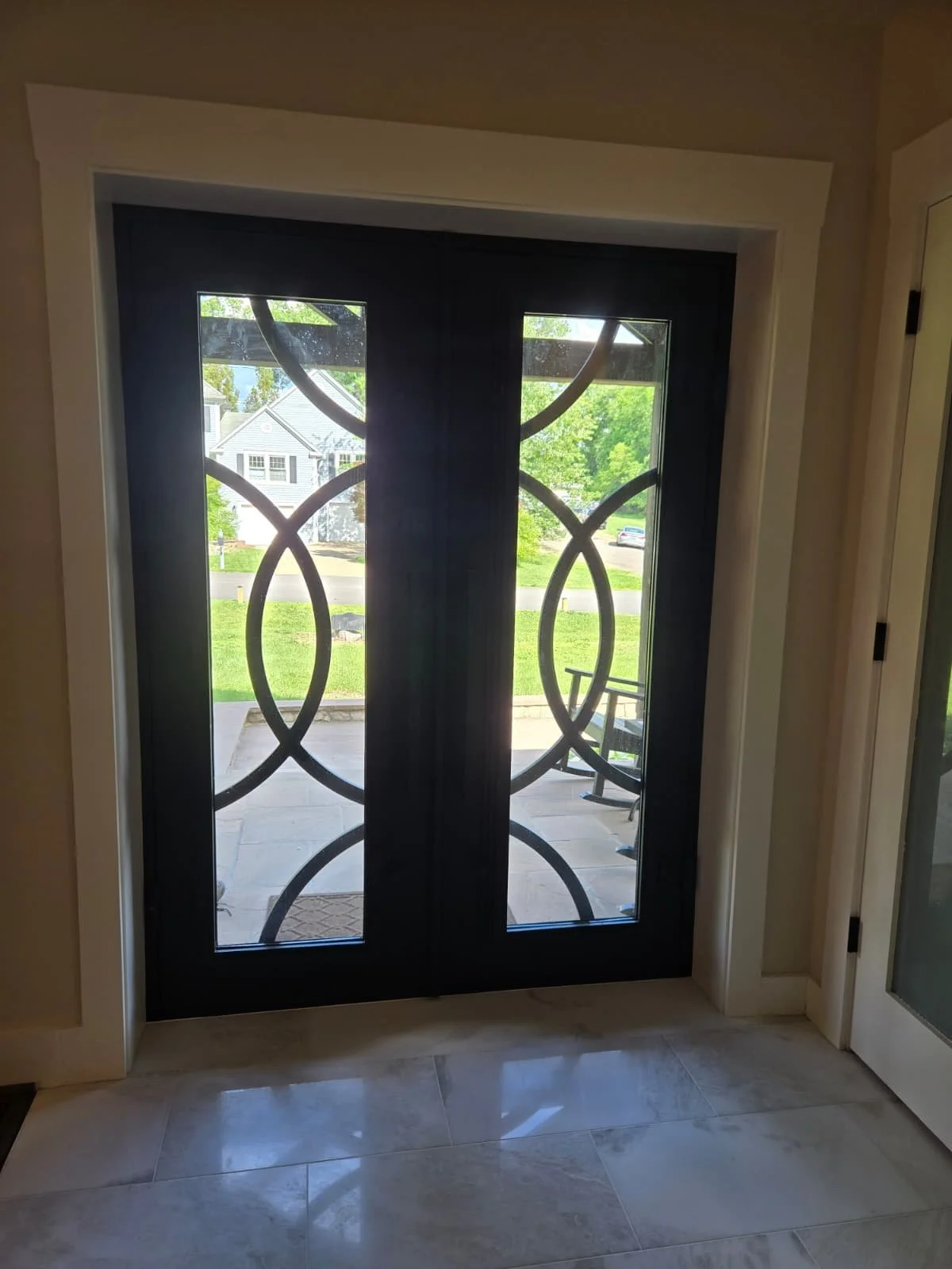 Black front door with glass panels and decorative metal scrollwork, leading to a porch with green lawn and trees visible outside.