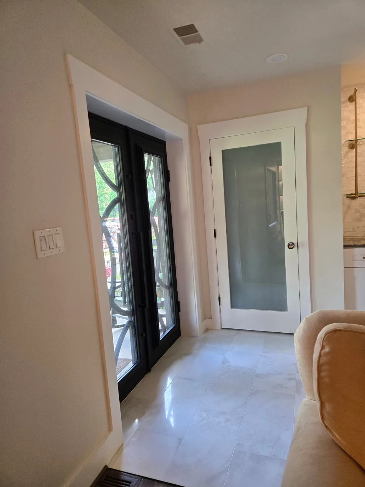 A corner of a living room with a beige armchair, a black-framed glass door, a white interior door with frosted glass, a wall-mounted light switch, and part of a kitchen area visible to the right.