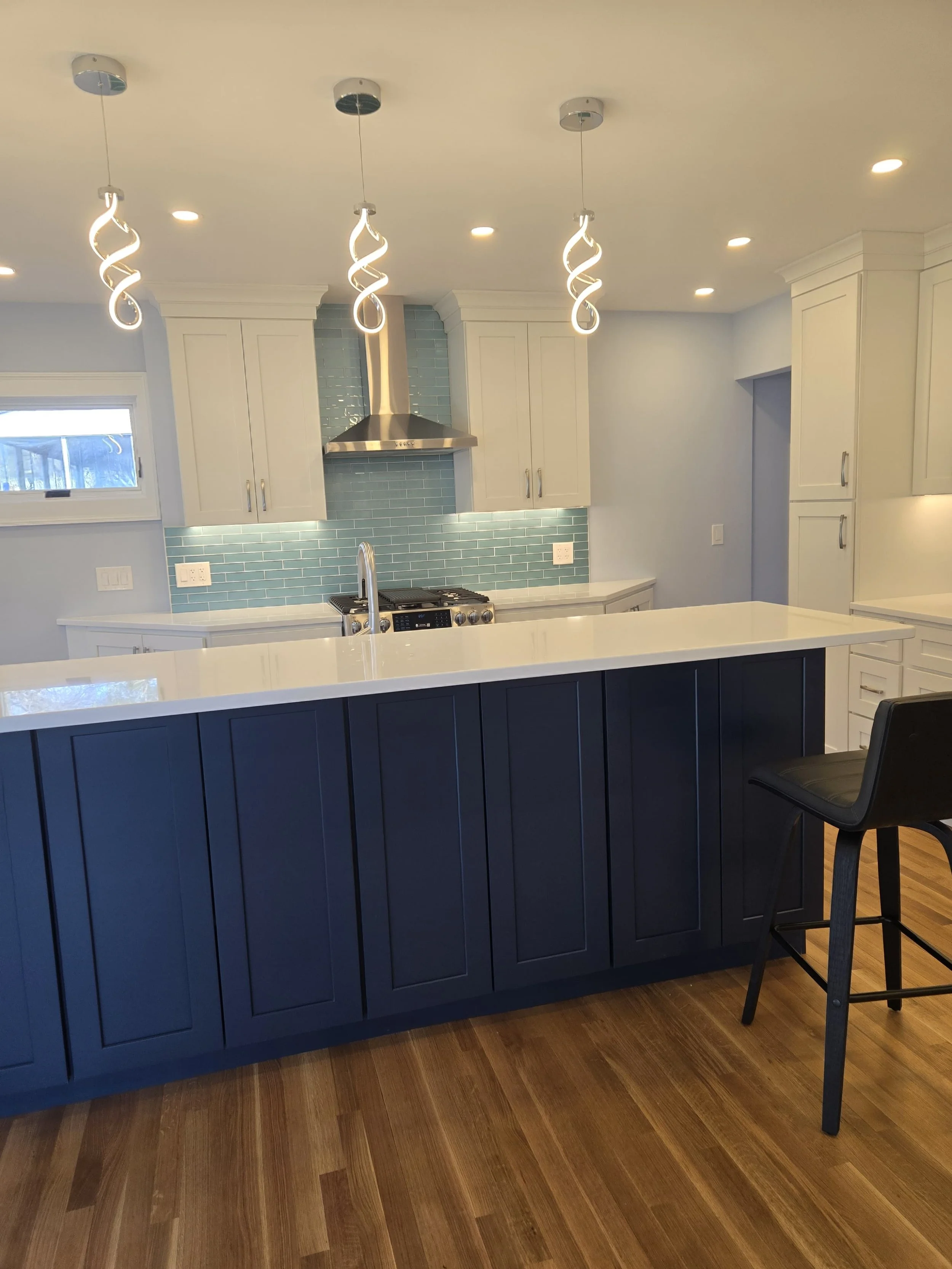 Modern kitchen with white cabinets, blue tiled backsplash, stainless steel range hood and oven, island with dark blue panels, and a black bar stool. Recessed ceiling lights and a small window are also visible.