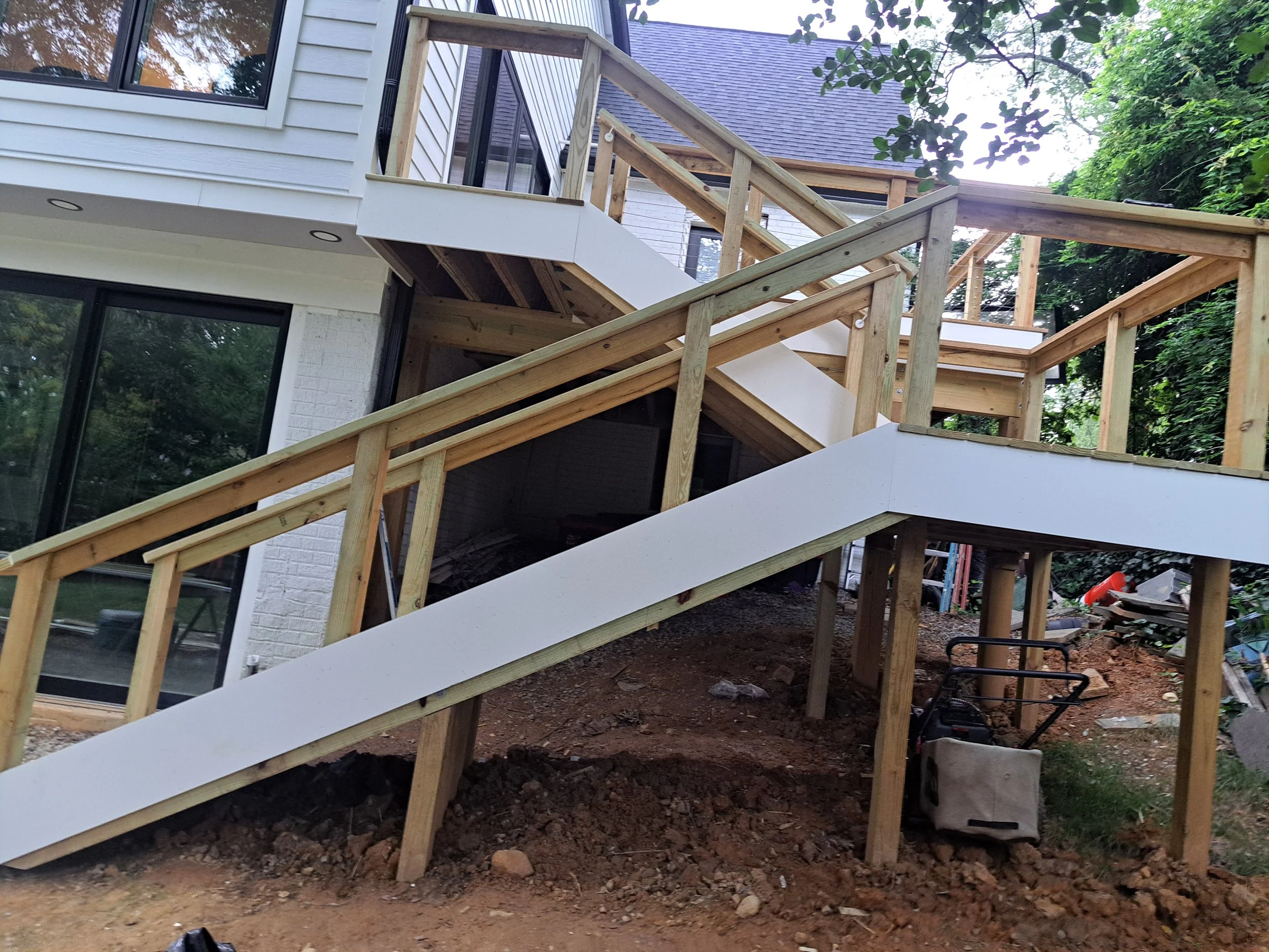 Residential deck construction shows a partially built deck with wooden framing and white stairs, attached to a house with white siding and large windows, surrounded by green trees.