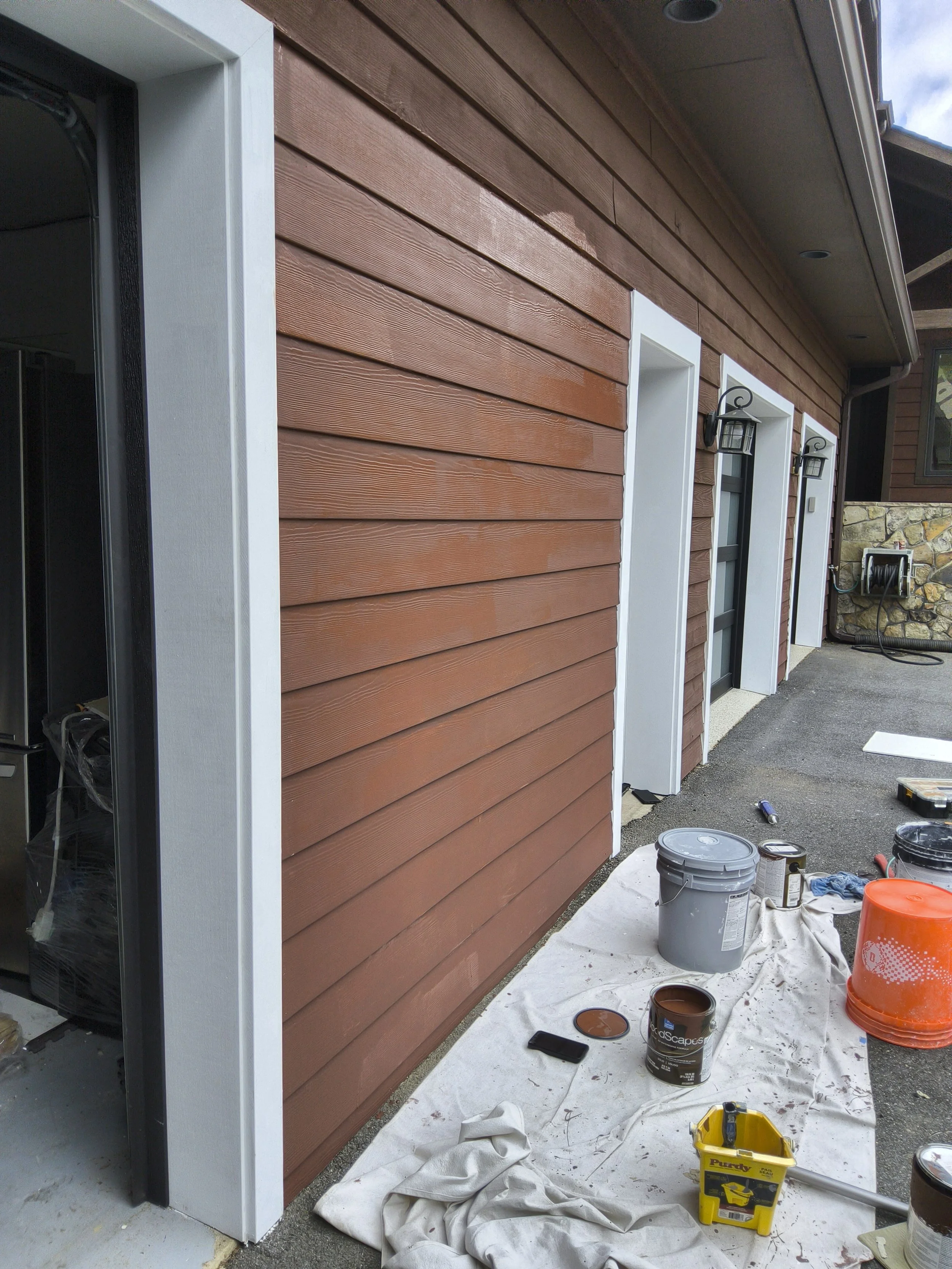 A house exterior with red wood siding, white window trims, black lamps, and renovation tools and supplies on a drop cloth, including paint cans, brushes, and a toolbox.