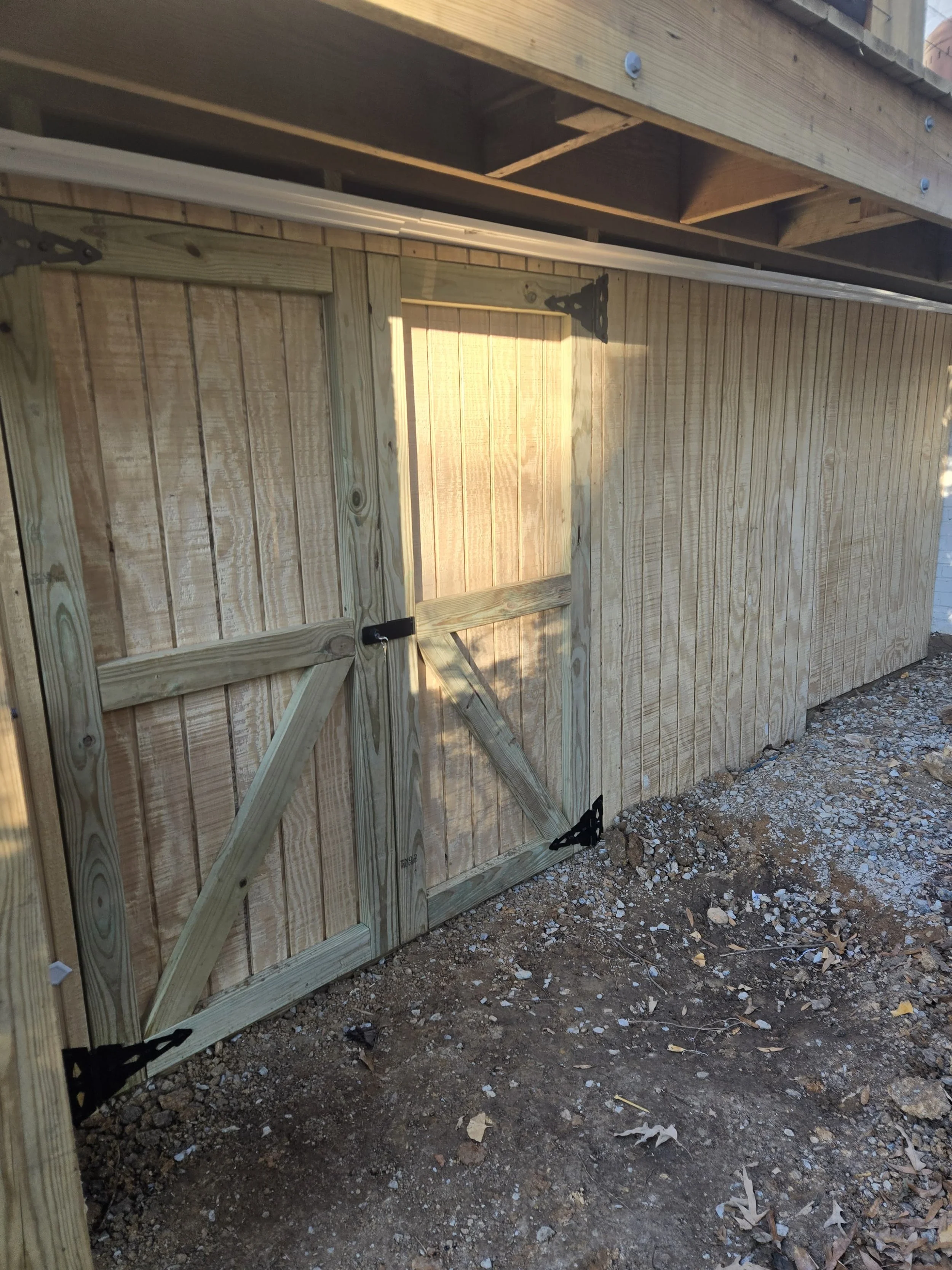 A newly built wooden backyard gate with a latch, attached to a wooden fence and a deck above, with dirt and small rocks on the ground.