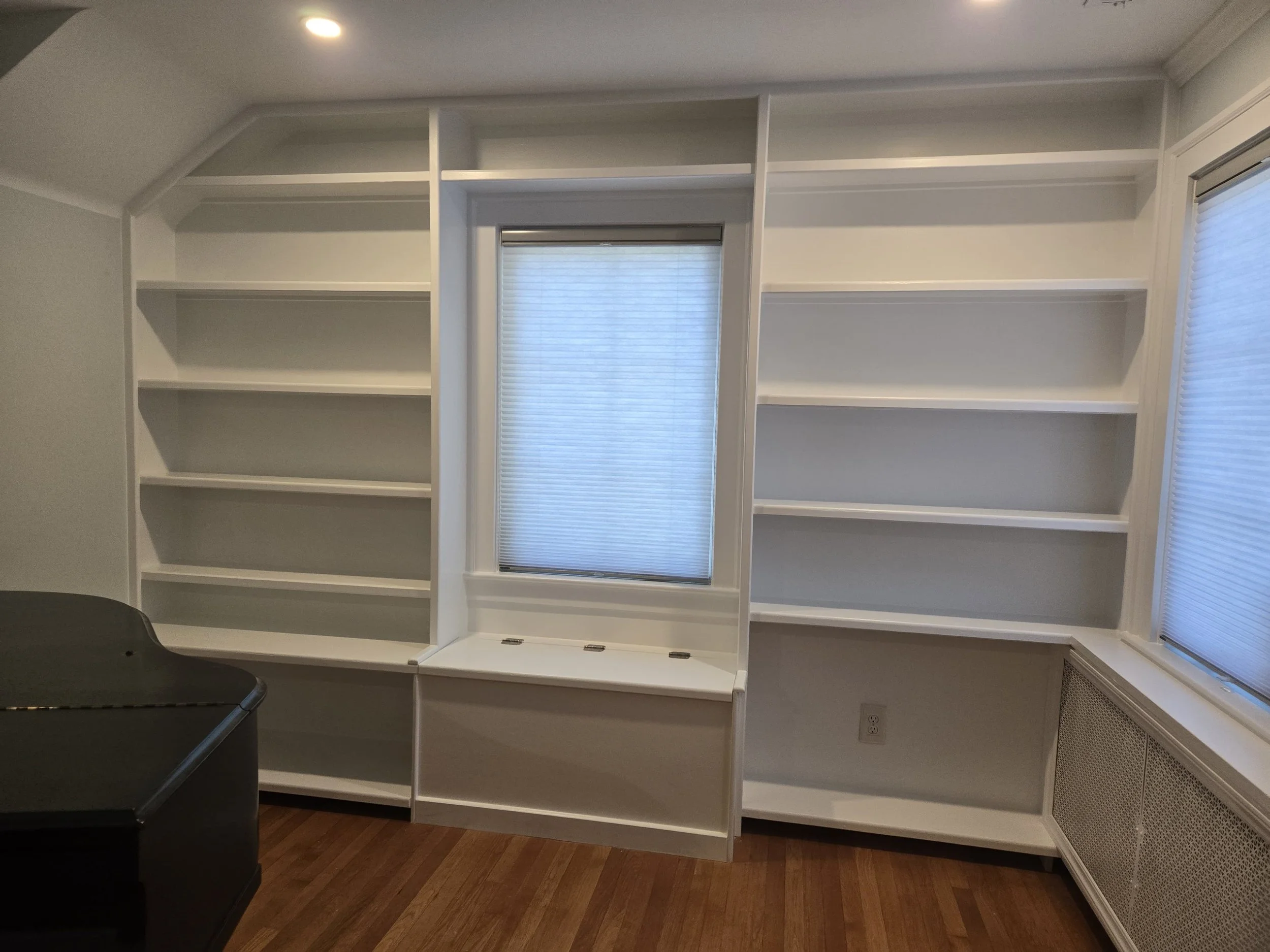 Empty white bookshelves in a room with a window and hardwood flooring.