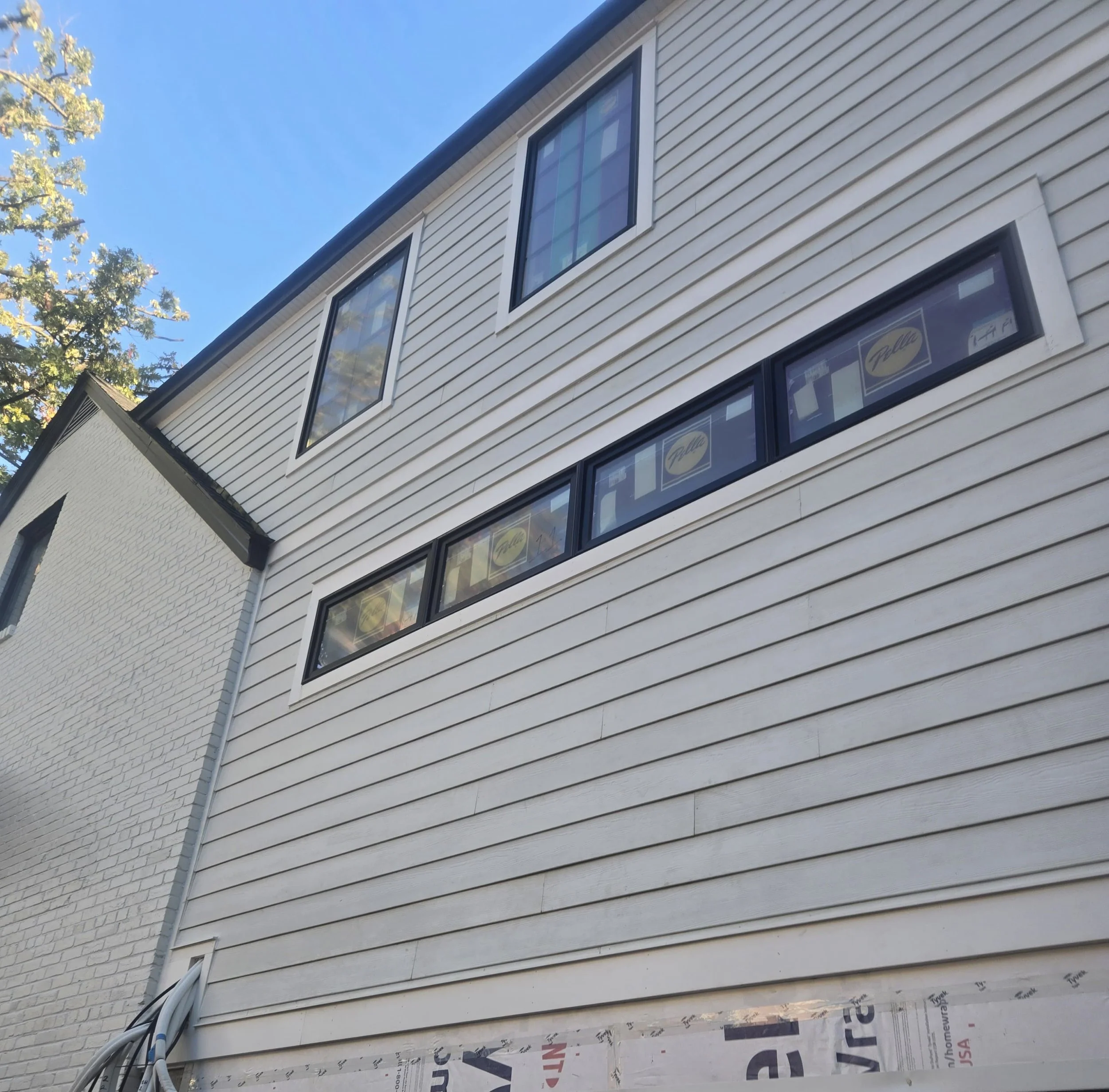 A close-up of a white house exterior with vinyl siding, large windows, and visible construction materials at the lower edge. A clear blue sky and tree branches are in the background.