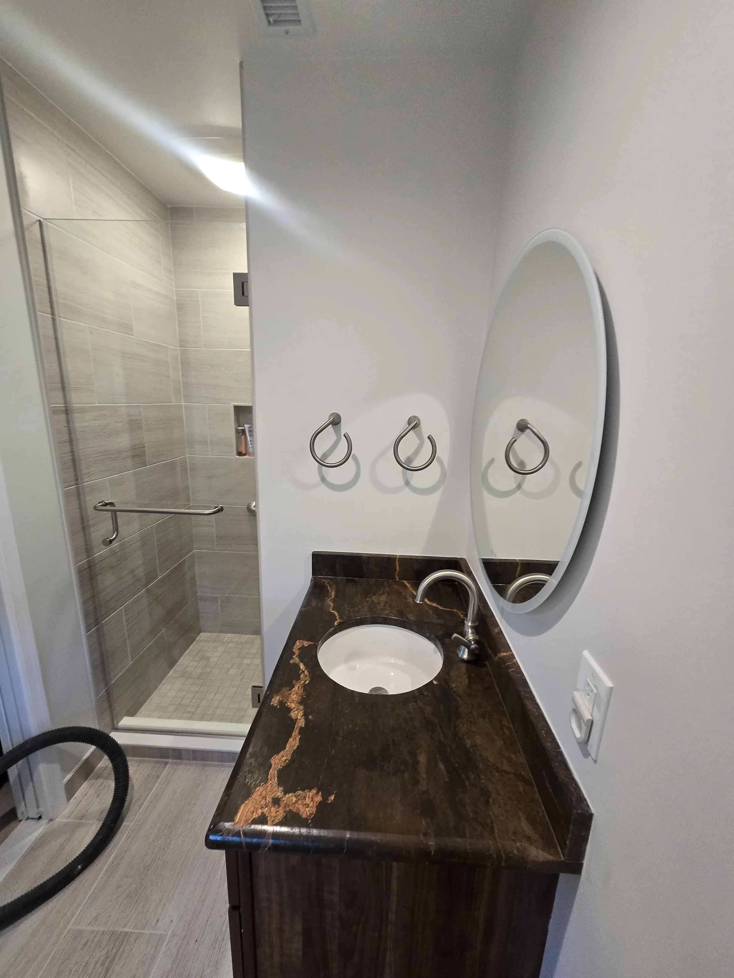Bathroom with a brown marble countertop, oval mirror, and a shower with a glass door and beige tiles.
