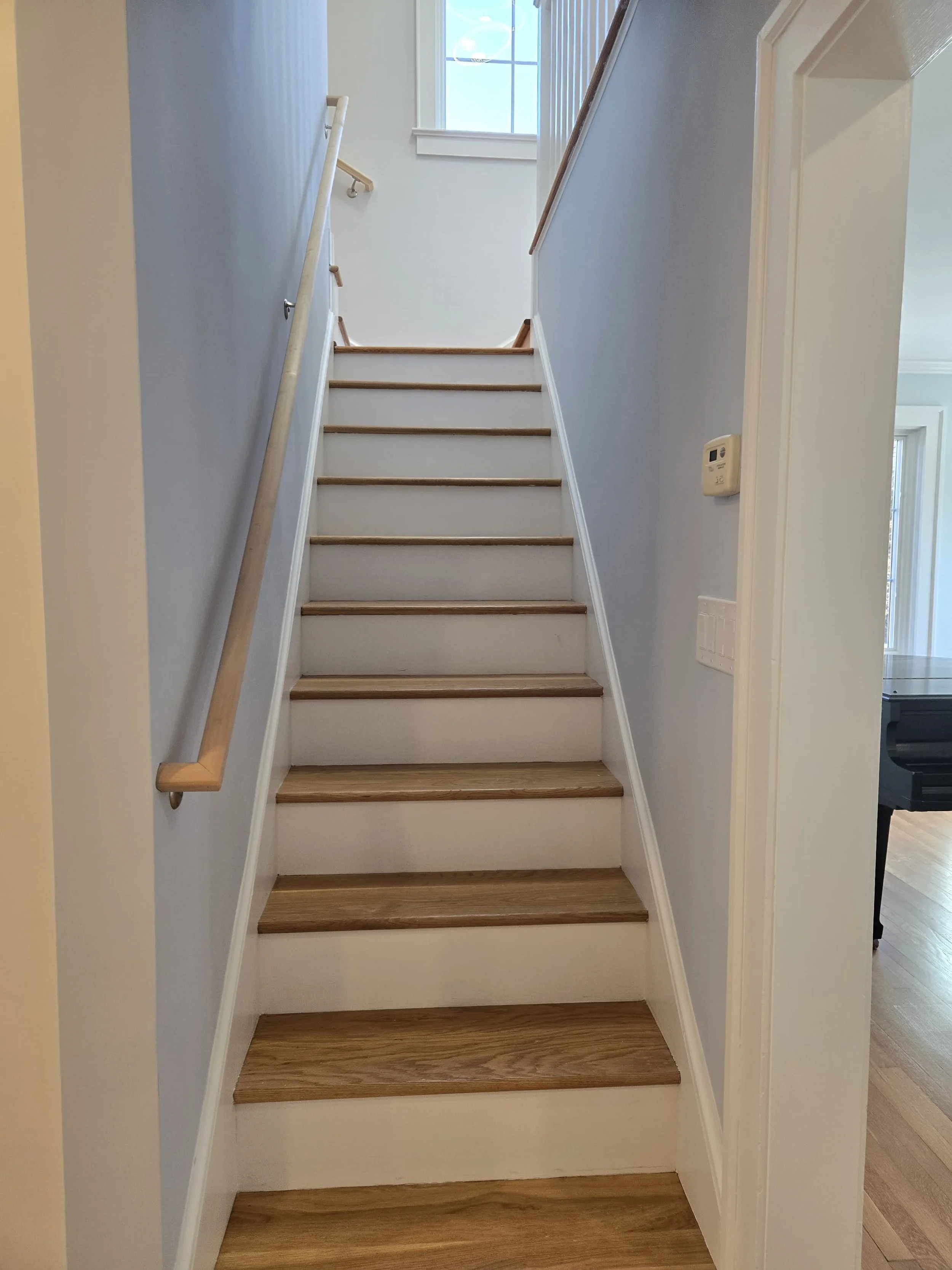 Interior view of a staircase with wooden steps, white walls, a wooden handrail on the left, and a window at the top providing natural light.