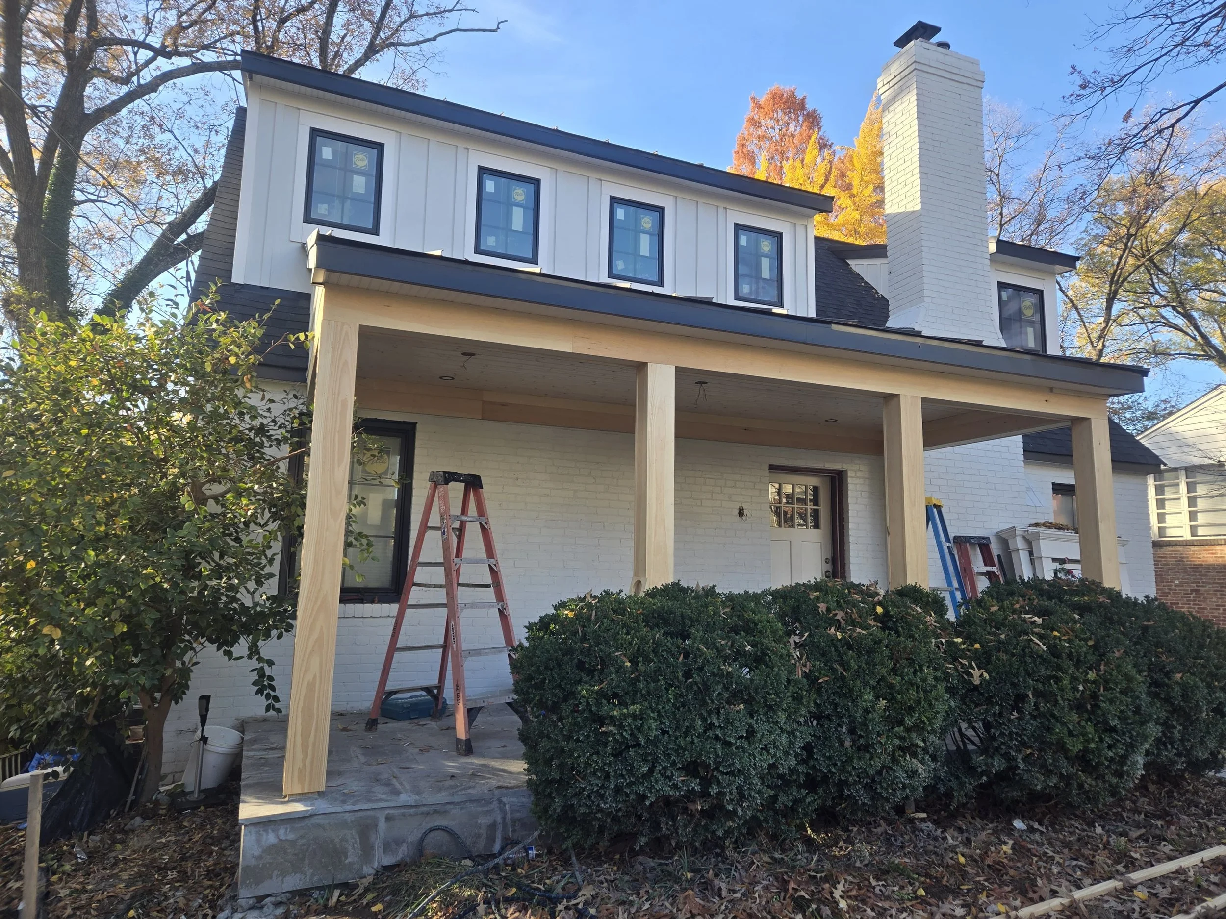 Construction site of a house with a partially built porch, ladders, and tools, surrounded by trees with fall foliage.