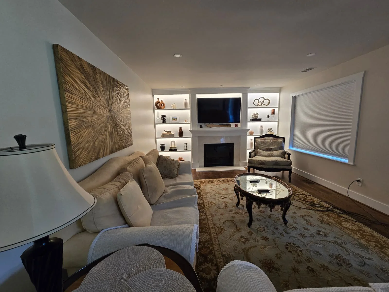 A cozy living room with a beige couch, a patterned armchair, a wooden coffee table, a fireplace with a mantle, built-in shelves with decorative items, a large wall art piece, a window with blinds, and a flat-screen TV.