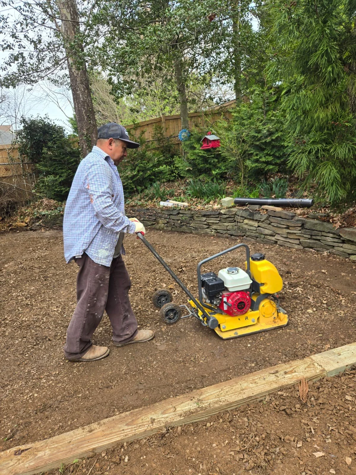 A man using a tabletop compactor to level a dirt yard in an outdoor garden with trees, shrubs, and a wooden fence in the background.