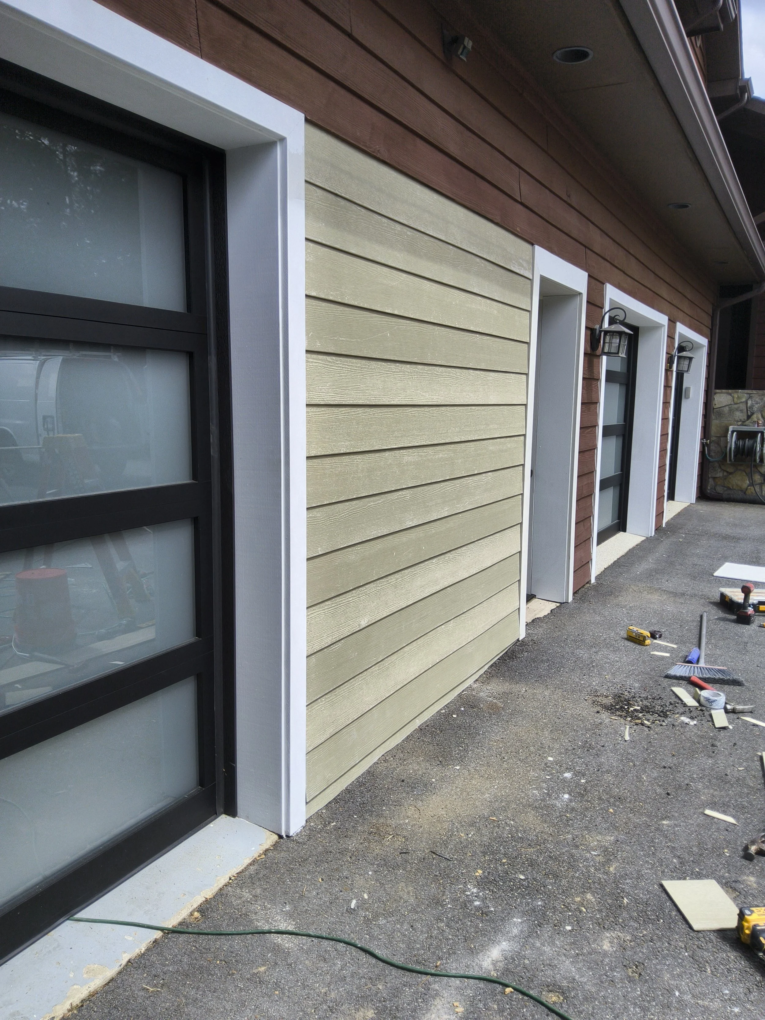 Exterior of a house under construction with beige siding, brown upper siding, black-framed windows, white trim, exterior lights, and construction tools on the ground.