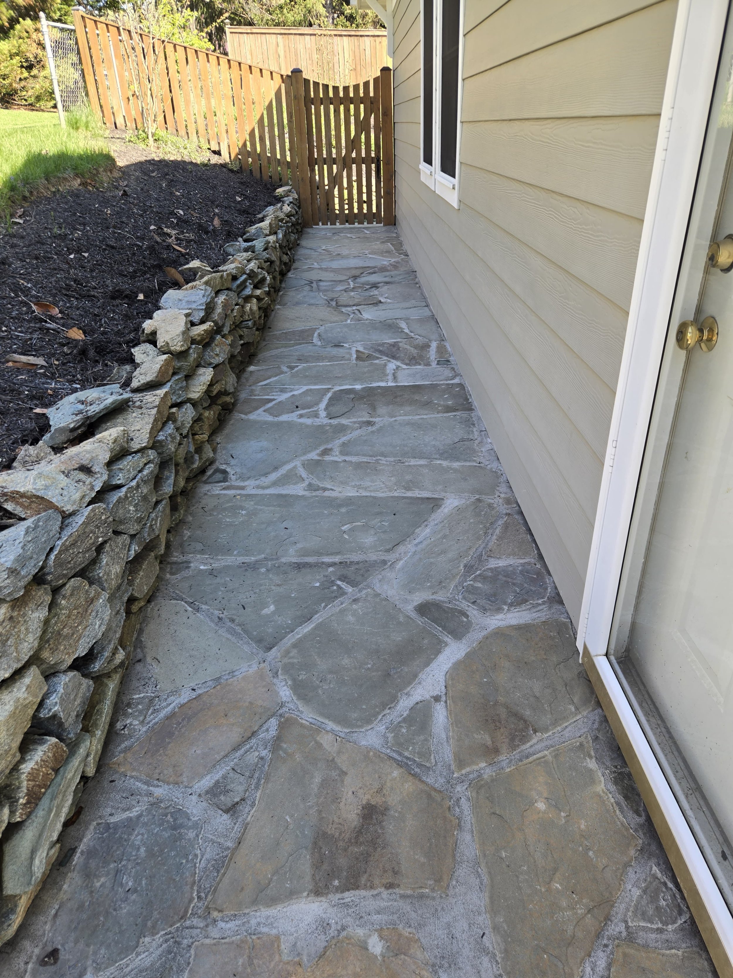 A narrow backyard pathway with stone flooring, a wood and stone retaining wall on the left, a beige house wall on the right, and a wooden gate at the end of the pathway.