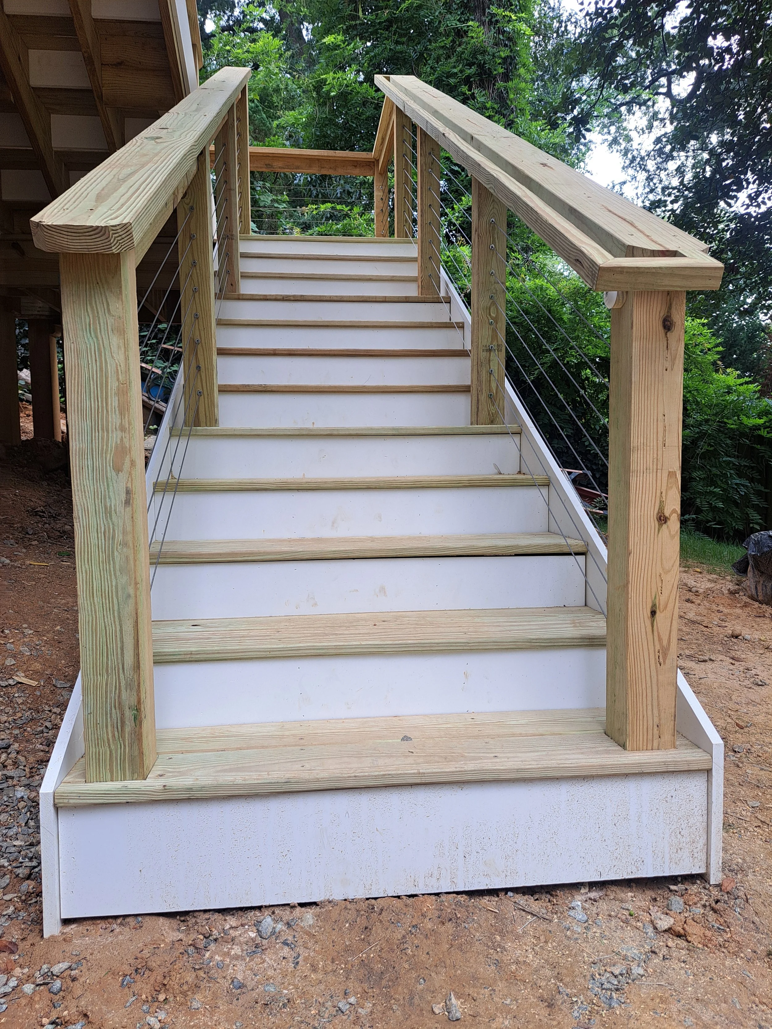 New wooden staircase outdoors with unfinished railing, leading upward, with green trees and sky in background, on dirt ground.