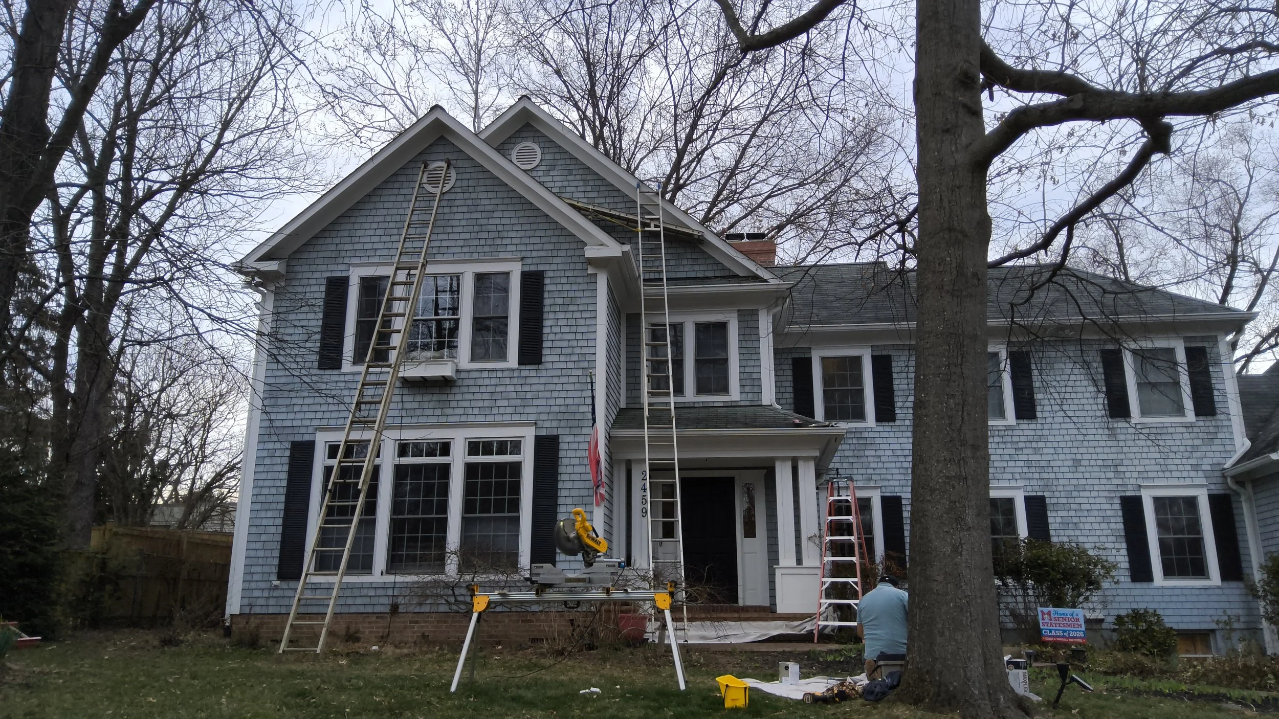 The exterior of a large blue-shingled house with multiple windows and black shutters is under repair. Two ladders are positioned against the house, and a work platform with a power saw is set in the driveway. A worker is visible near the base of a large tree, and a sign indicates a senior class of 2024. Bare trees suggest it is winter or late fall.