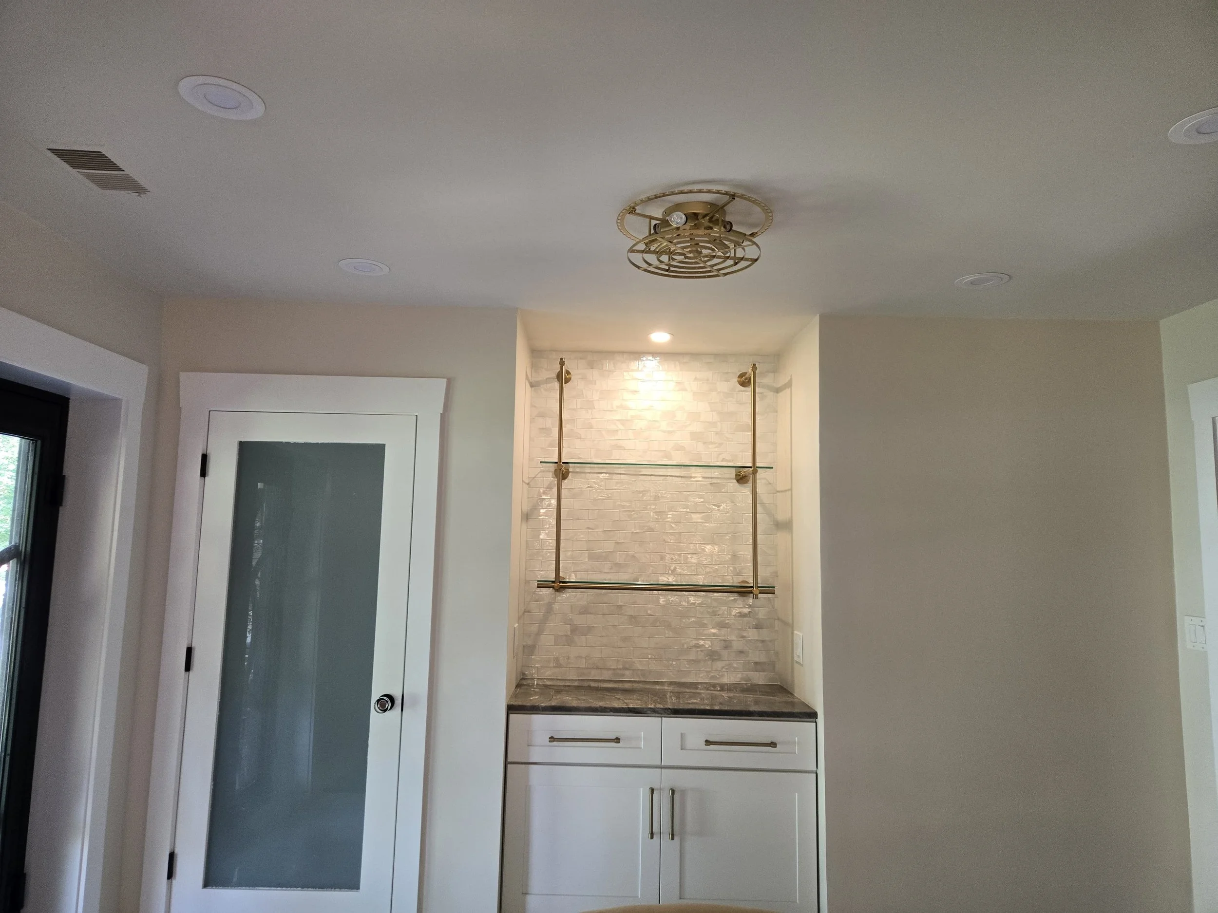 The image shows a room with a white ceiling, a modern chandelier, a door with frosted glass, a marble countertop, and white cabinets with gold handles.