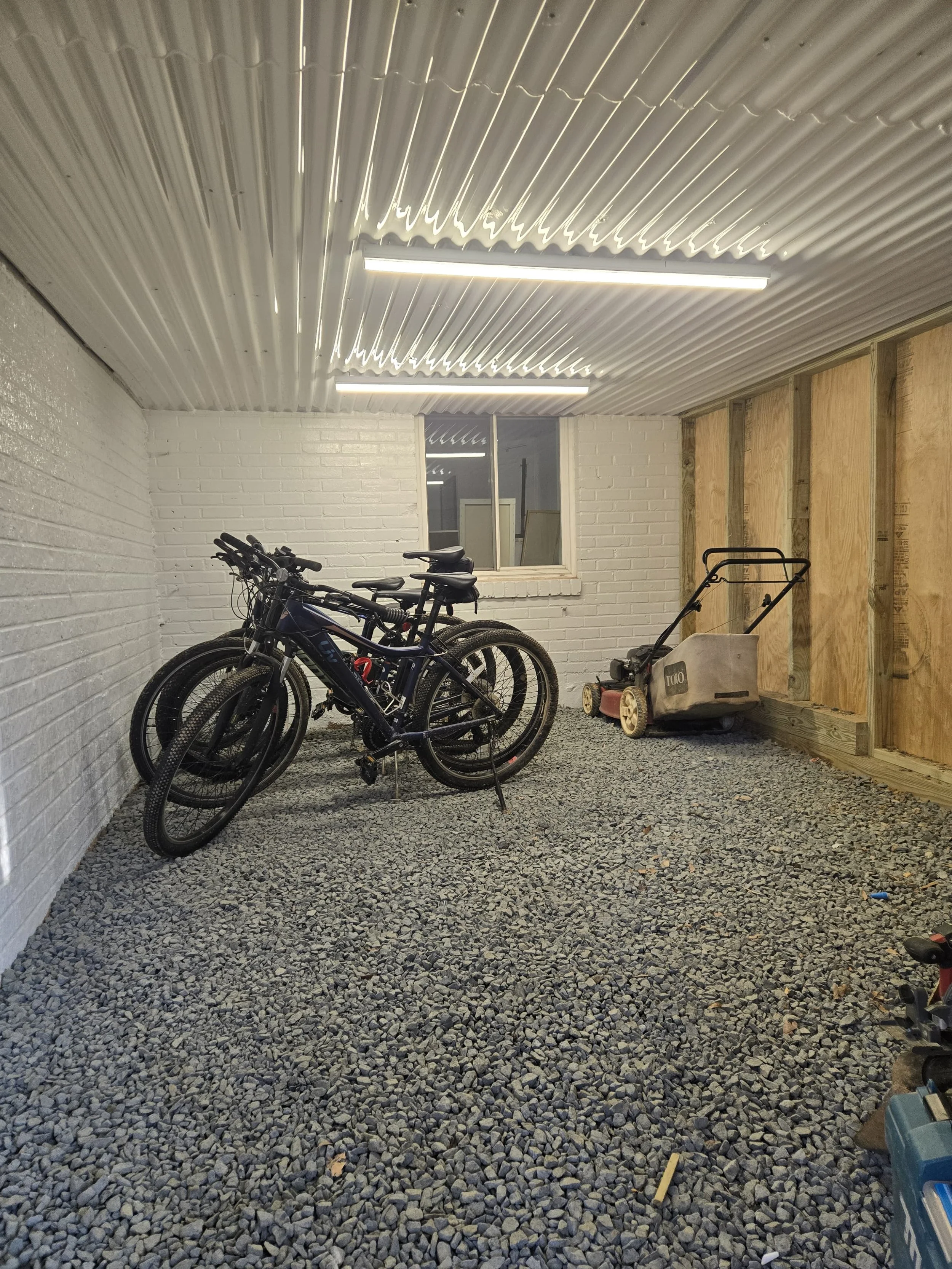 Indoor space with a corrugated metal ceiling, fluorescent lights, a white painted brick wall, a window, and construction equipment including two bicycles, a lawnmower, and various tools.