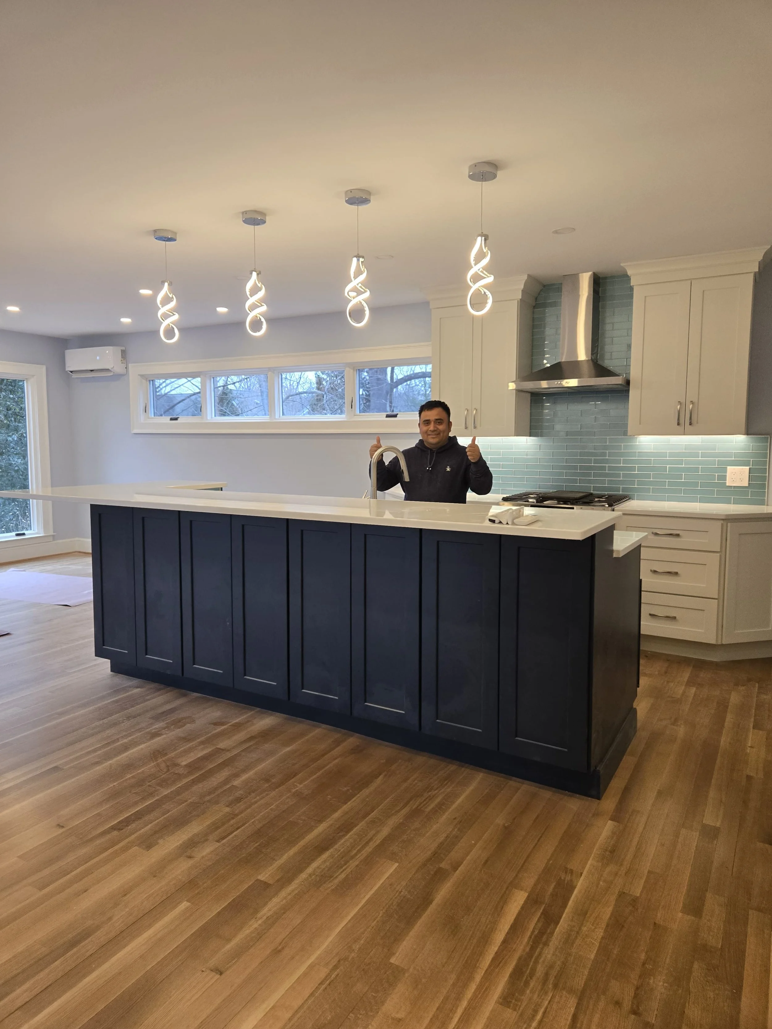 A man standing behind a kitchen island with a sink, giving a thumbs up, in a modern kitchen with white cabinets, stainless steel range hood, teal subway tile backsplash, and wood flooring.