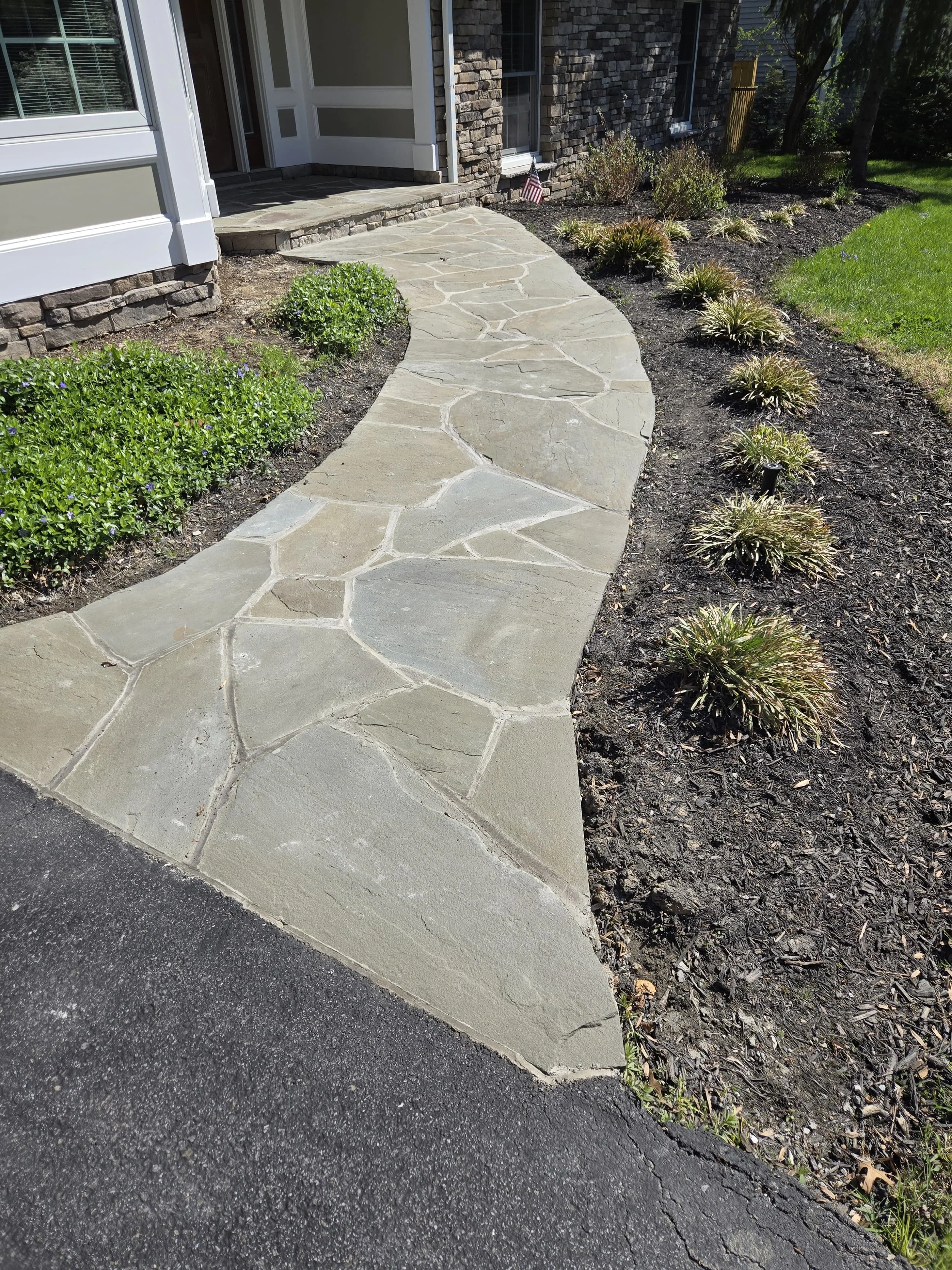 A curved stone walkway leading to the front door of a house, bordered by flower beds with green shrubs and plants, with a mix of mulch and grass in a suburban yard.
