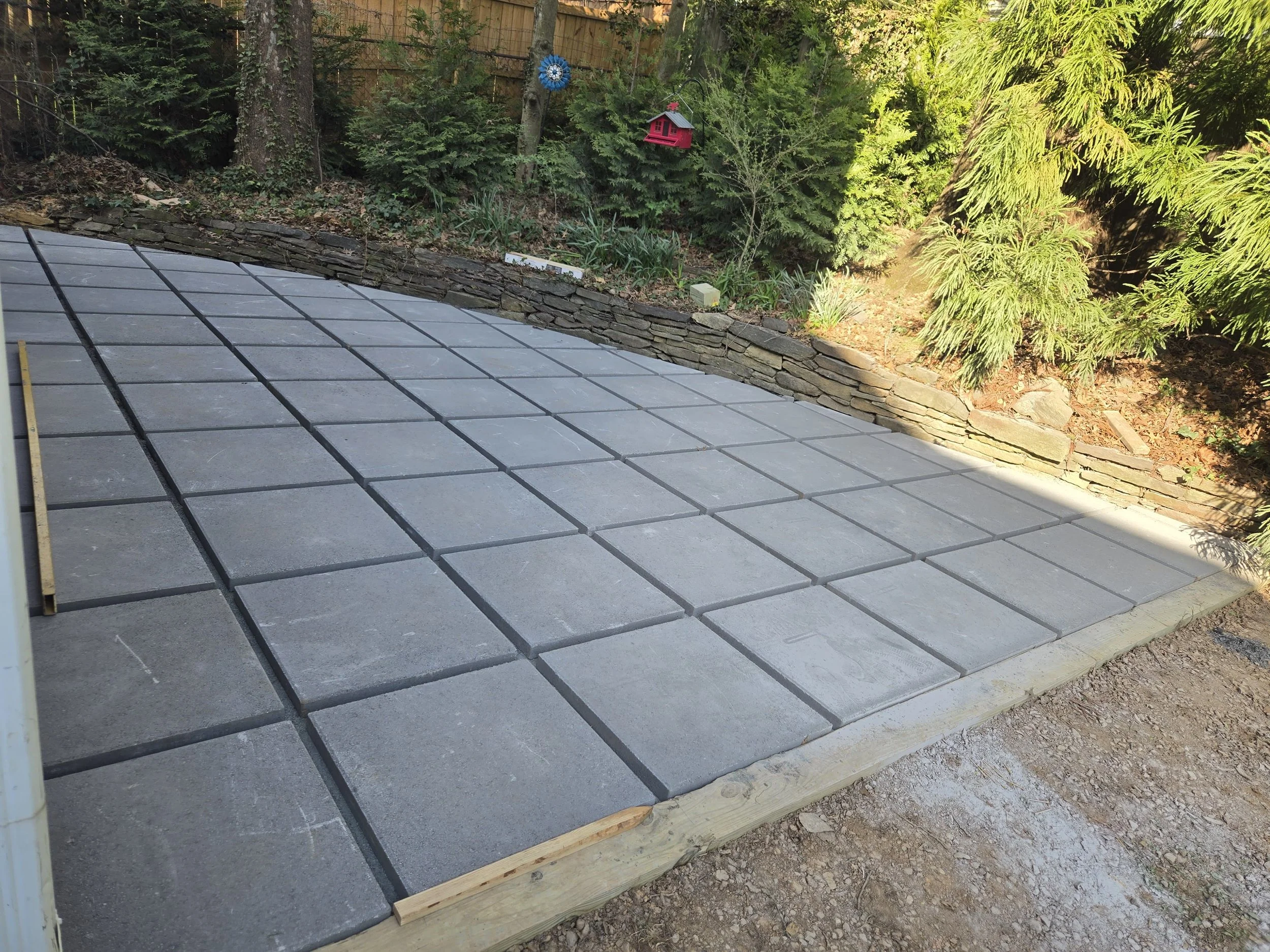 A backyard with a partially completed patio made of gray pavers, bordered by a curved stone retaining wall. Behind the wall, there are trees and shrubs, with decorative birdhouses hanging from the branches.