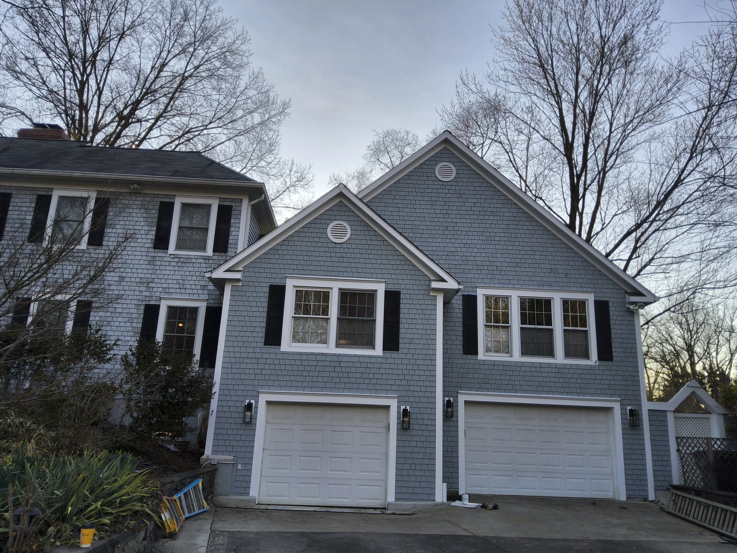 Front of a two-story house with blue shingle siding, black shutters, multiple windows, a garage door, and a sloped roof with round vents, on a clear day with leafless trees in the background.