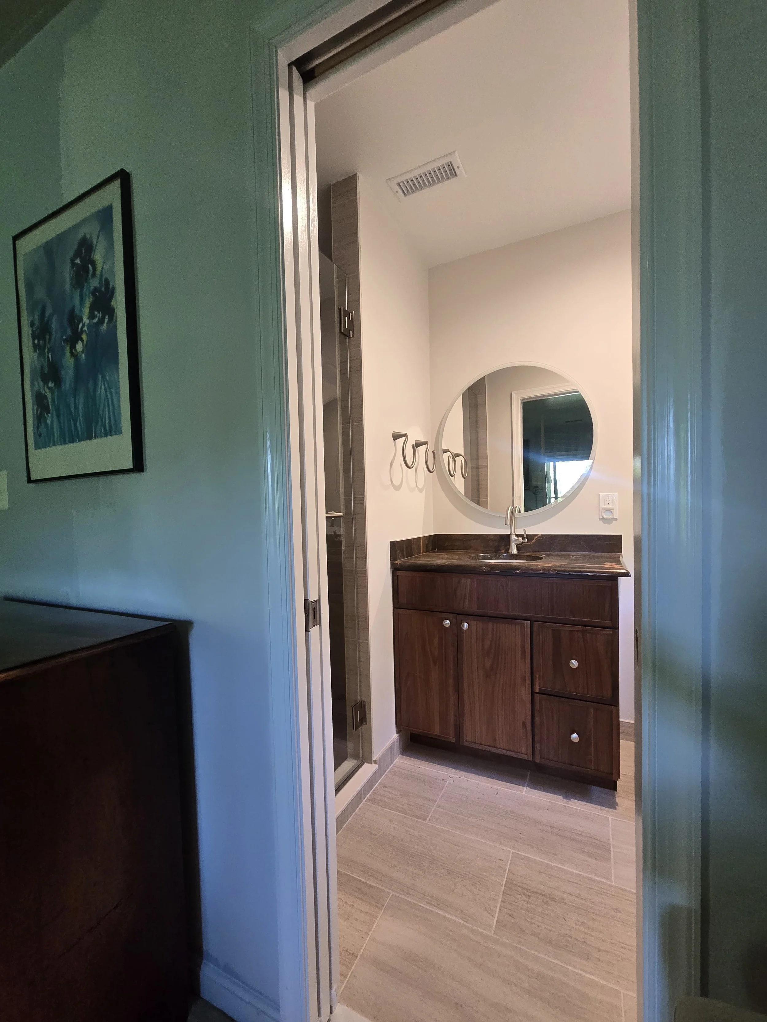 Bathroom with wooden cabinet, round mirror, towel holders, and a shower door, with beige tile flooring and white walls.