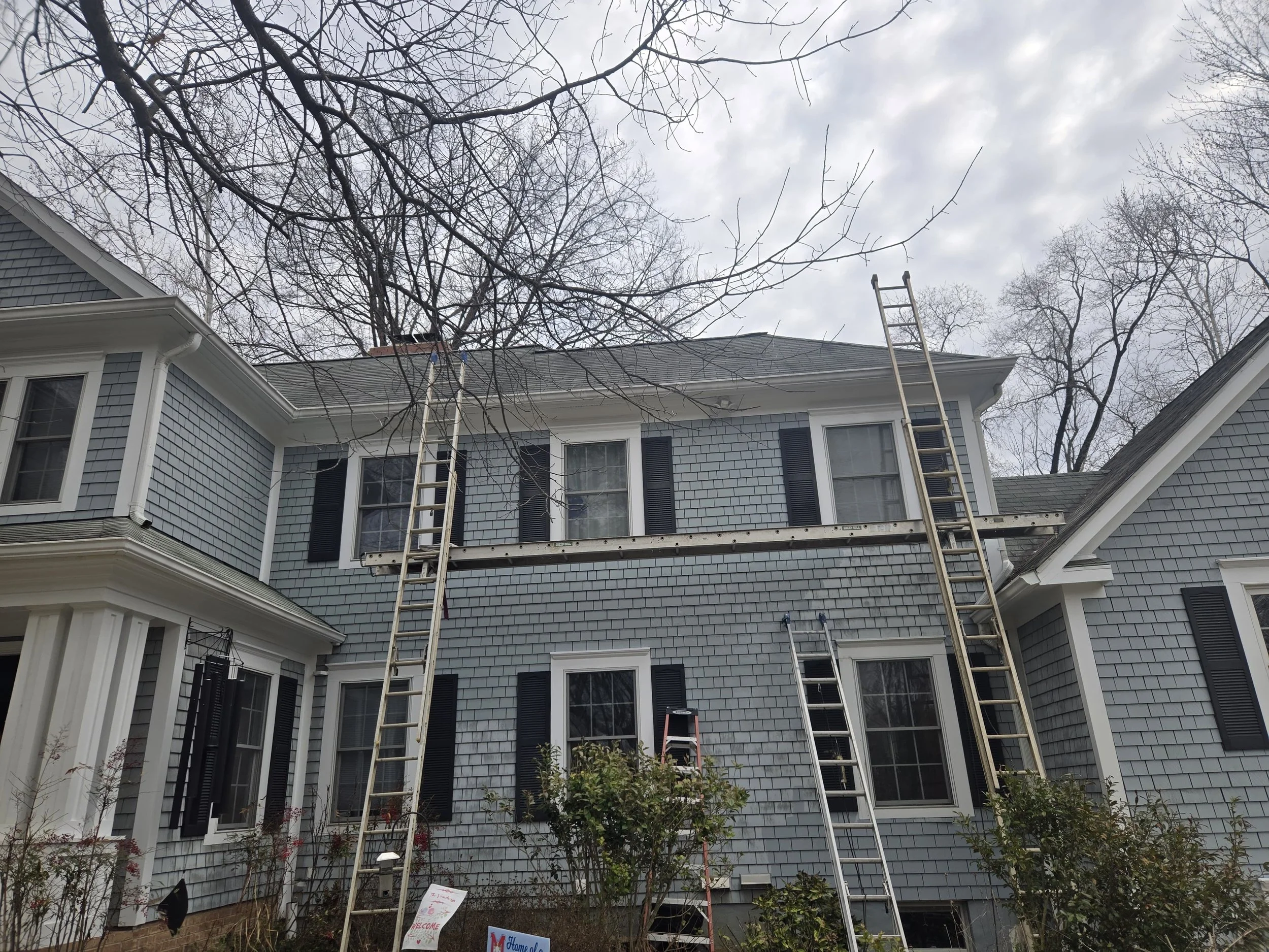 The image shows the exterior of a light blue, multi-story house with black shutters and white trim. There are climbing ladders against the house, reaching the roof. The sky is overcast and several leafless trees are visible around the house, indicating it is likely late fall or winter.