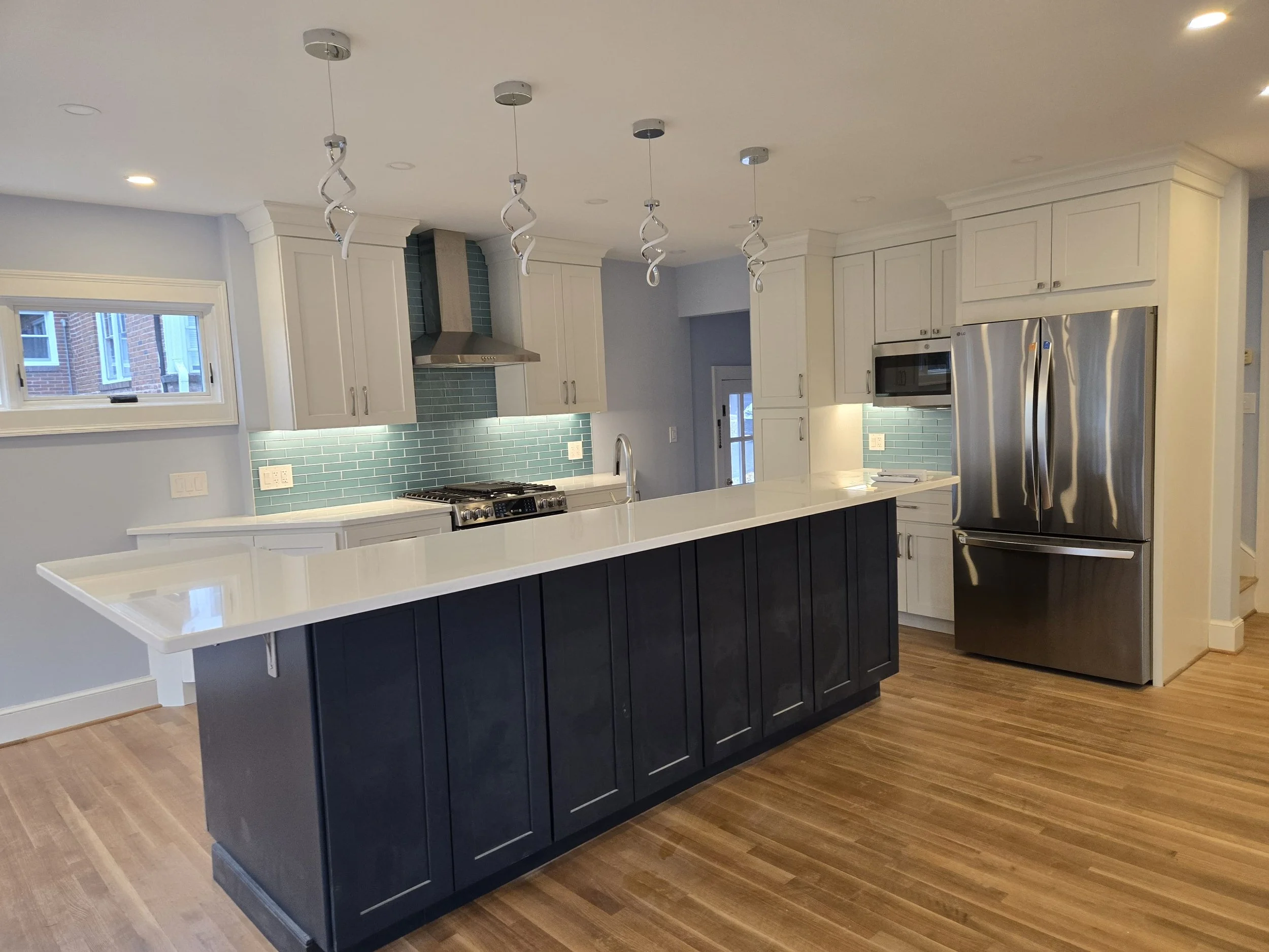 Modern kitchen with white cabinets, green tile backsplash, stainless steel appliances, and a dark blue kitchen island with a white countertop. There are pendant lights hanging from the ceiling and a window with a view of neighboring brick buildings.