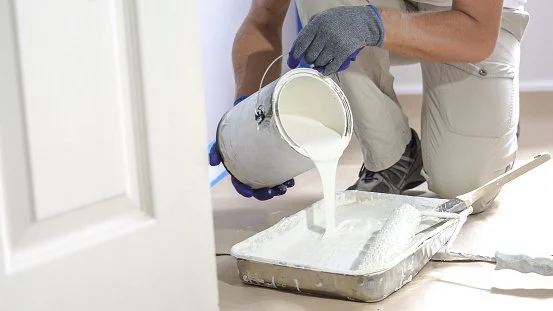 A person pouring white paint into a tray with brush, wearing gloves and kneeling on the floor.