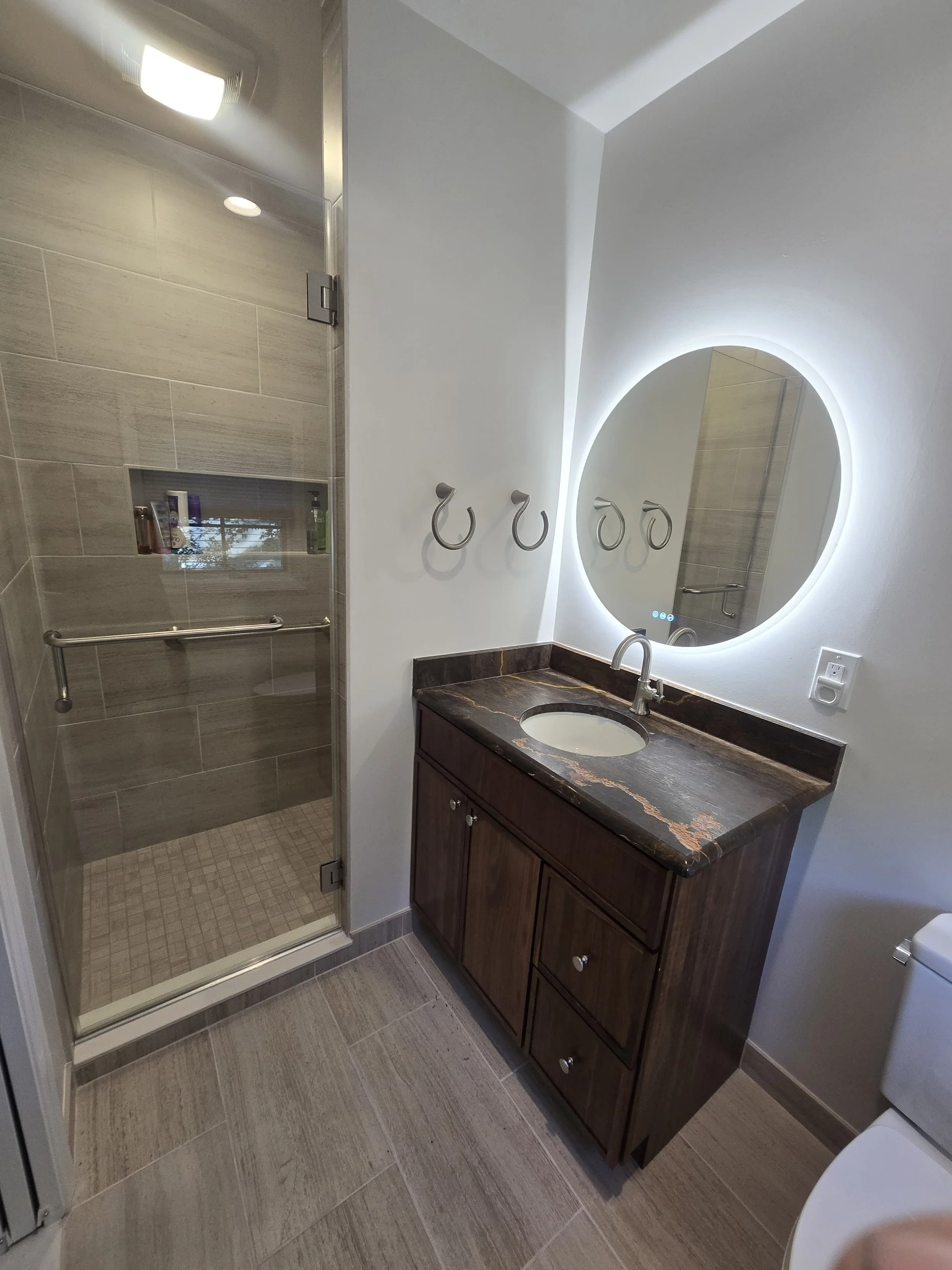 Bathroom with a walk-in shower, wooden vanity with a granite countertop, oval mirror with LED backlight, towel hooks, and a toilet.