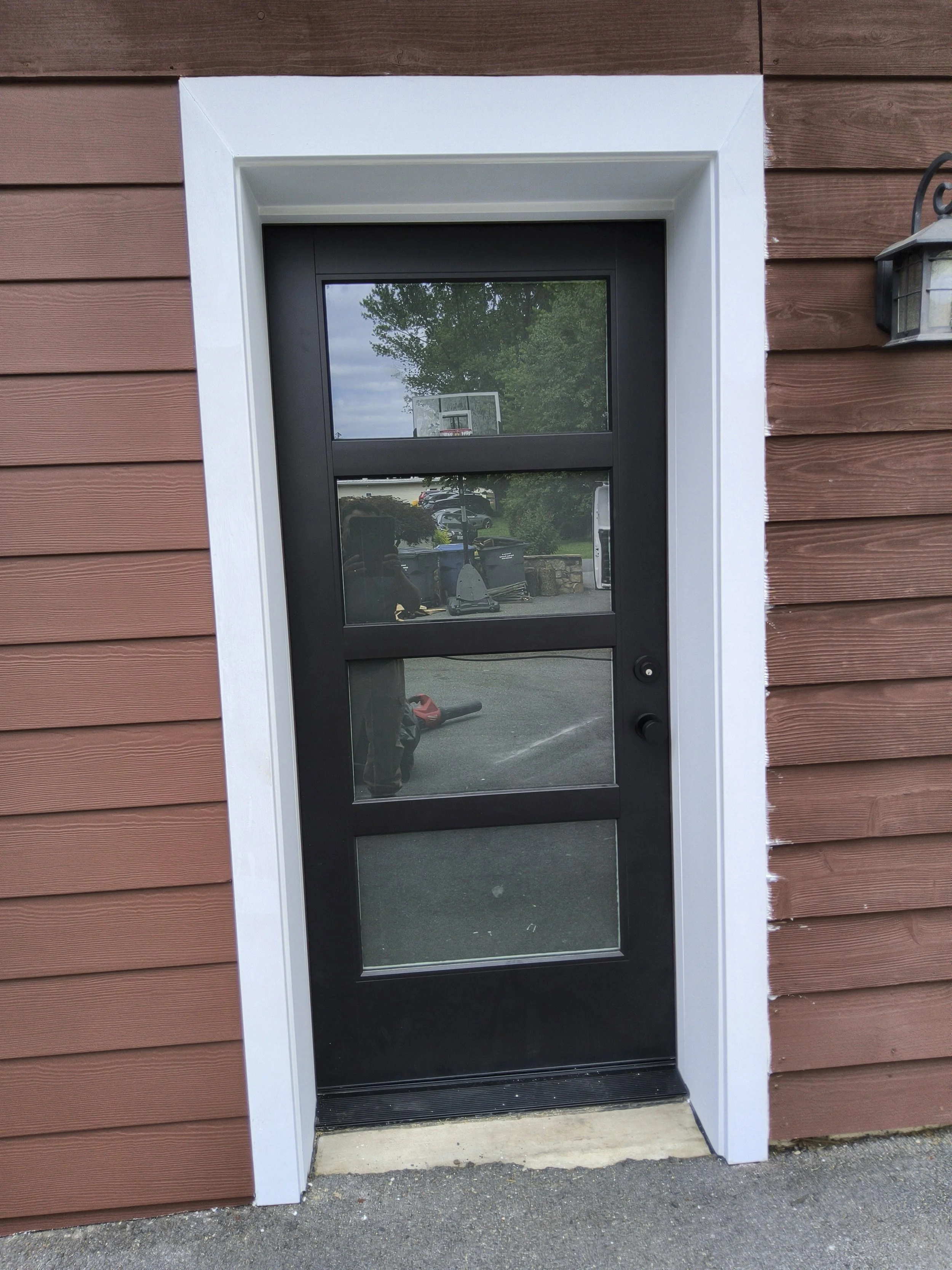 Close-up of a black-framed glass door on a red wooden building, with a white trim and a small step at the threshold. Reflection in the glass shows a person taking the photo, trees, a basketball hoop, parked cars, and garbage bins outside.