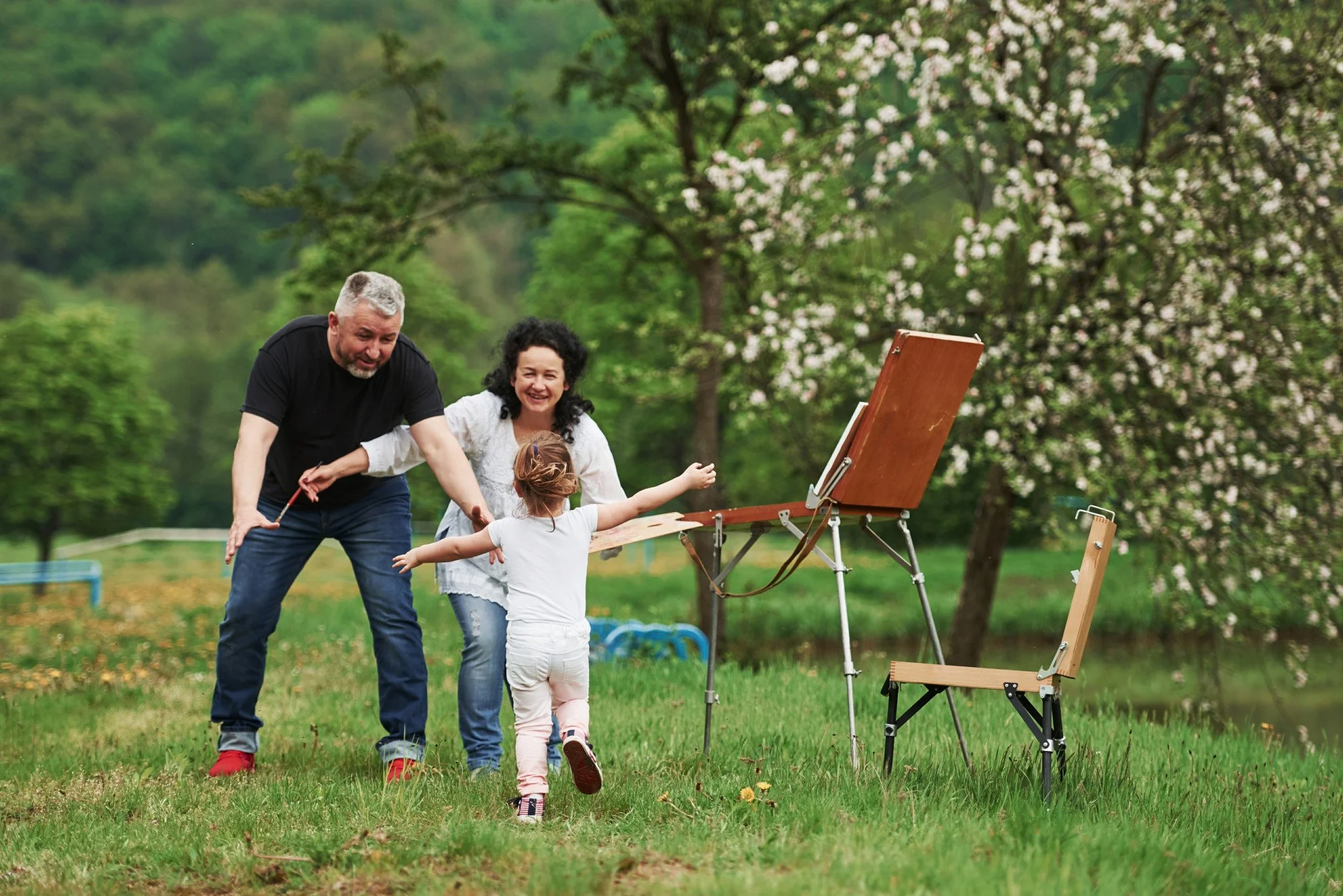 A family of three, two adults and a child, enjoying their time outdoors on a sunny day in a park with green grass and blooming trees. The children appears to be running towards a large wooden artist's easel set up on a portable stand, while the adults cheer and interact with the child. The scene exudes happiness and togetherness amidst a lush natural setting.