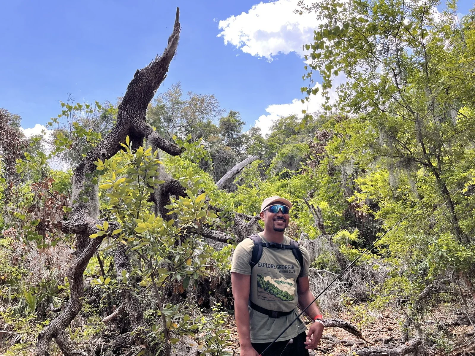 A man wearing a gray t-shirt, tan hat, blue sunglasses, and a backpack, smiling while hiking in a forested area with a fallen tree and lush green foliage under a partly cloudy sky.