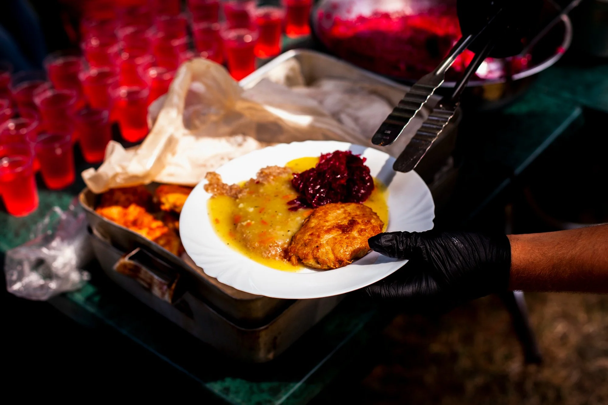 A person wearing black gloves holds a white plate with a piece of cooked fish, beaten eggs, and red berry compote. In the background, there is a buffet with several small cups filled with red liquid.