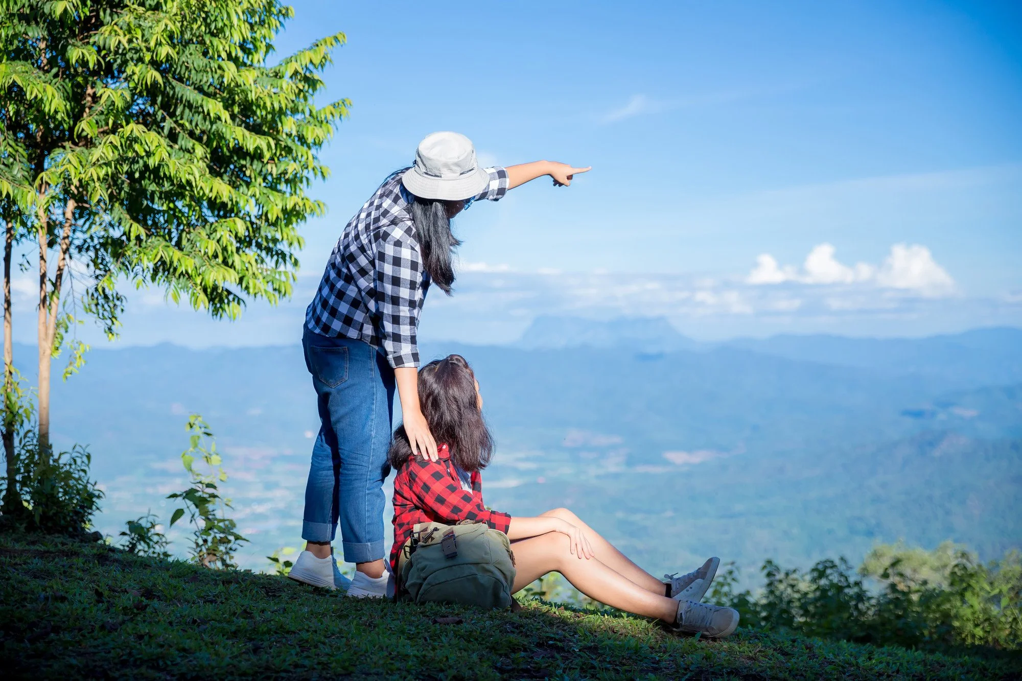 A woman, a young girl, and a woman in a hat are outdoors on a grassy hill. The woman with a hat and black-and-white checkered shirt is pointing at the sky. The girl, sitting and wearing a red and black checkered shirt, is looking in the same direction. The background features a clear blue sky with some clouds, a tree, and distant mountains.