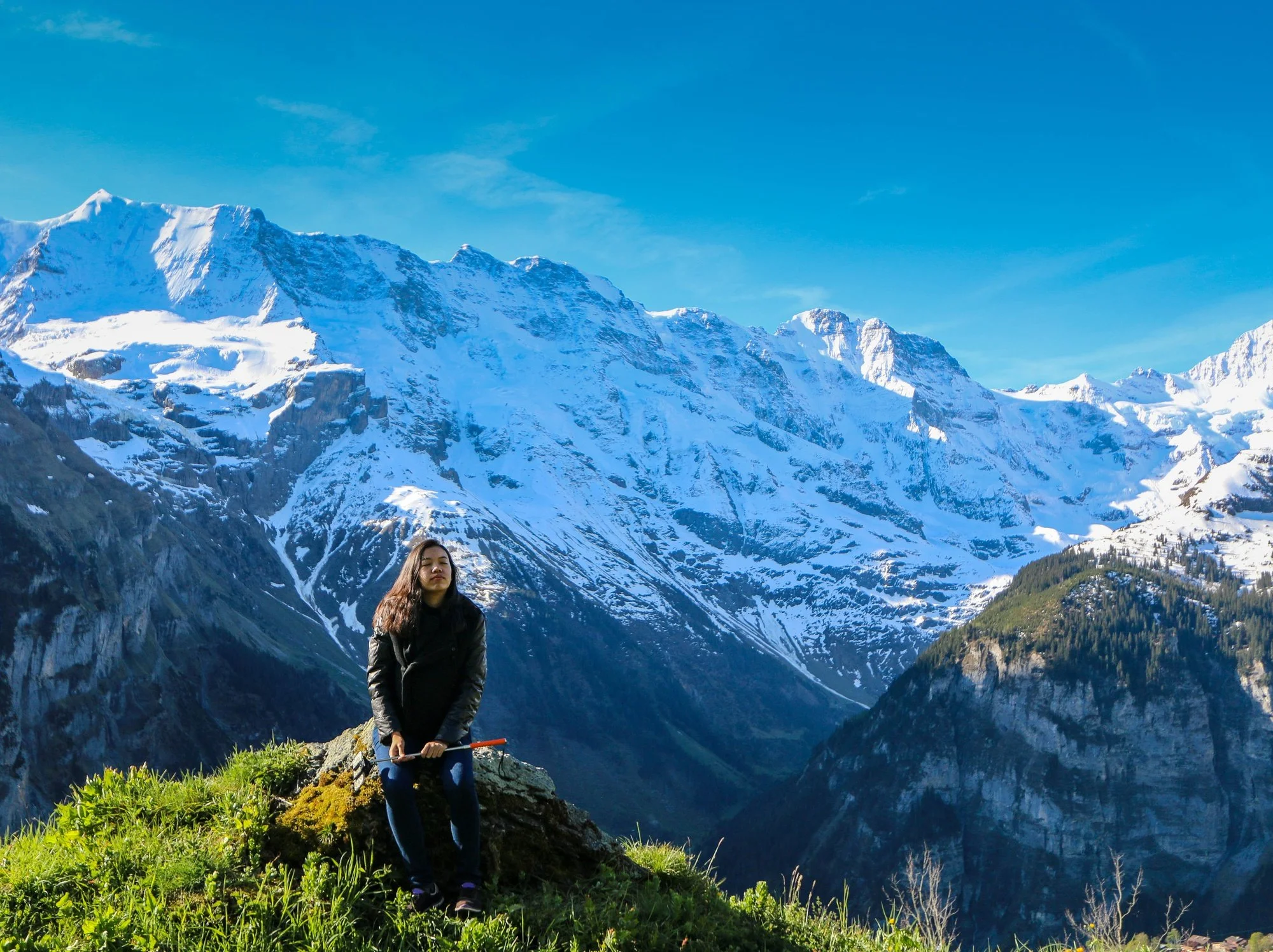 A woman standing on a grassy ledge with a snow-capped mountain range in the background on a clear day.