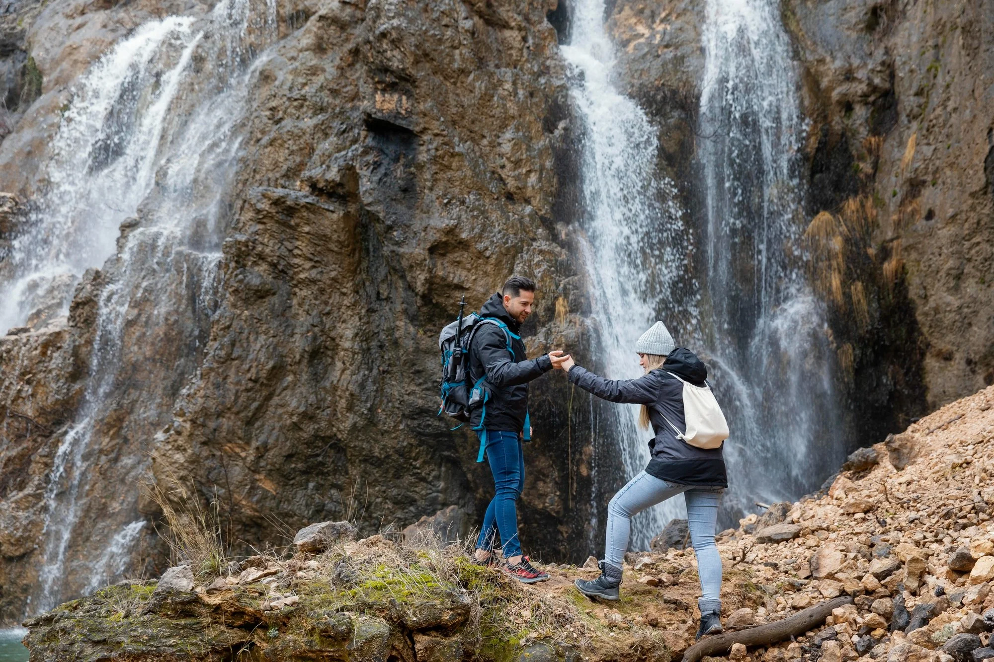Two hikers, one man and one woman, near a waterfall with rugged rocks and sparse vegetation. The man is wearing a dark jacket and the woman is wearing a gray beanie and gray jacket. The woman is helping the man step over a rocky patch.
