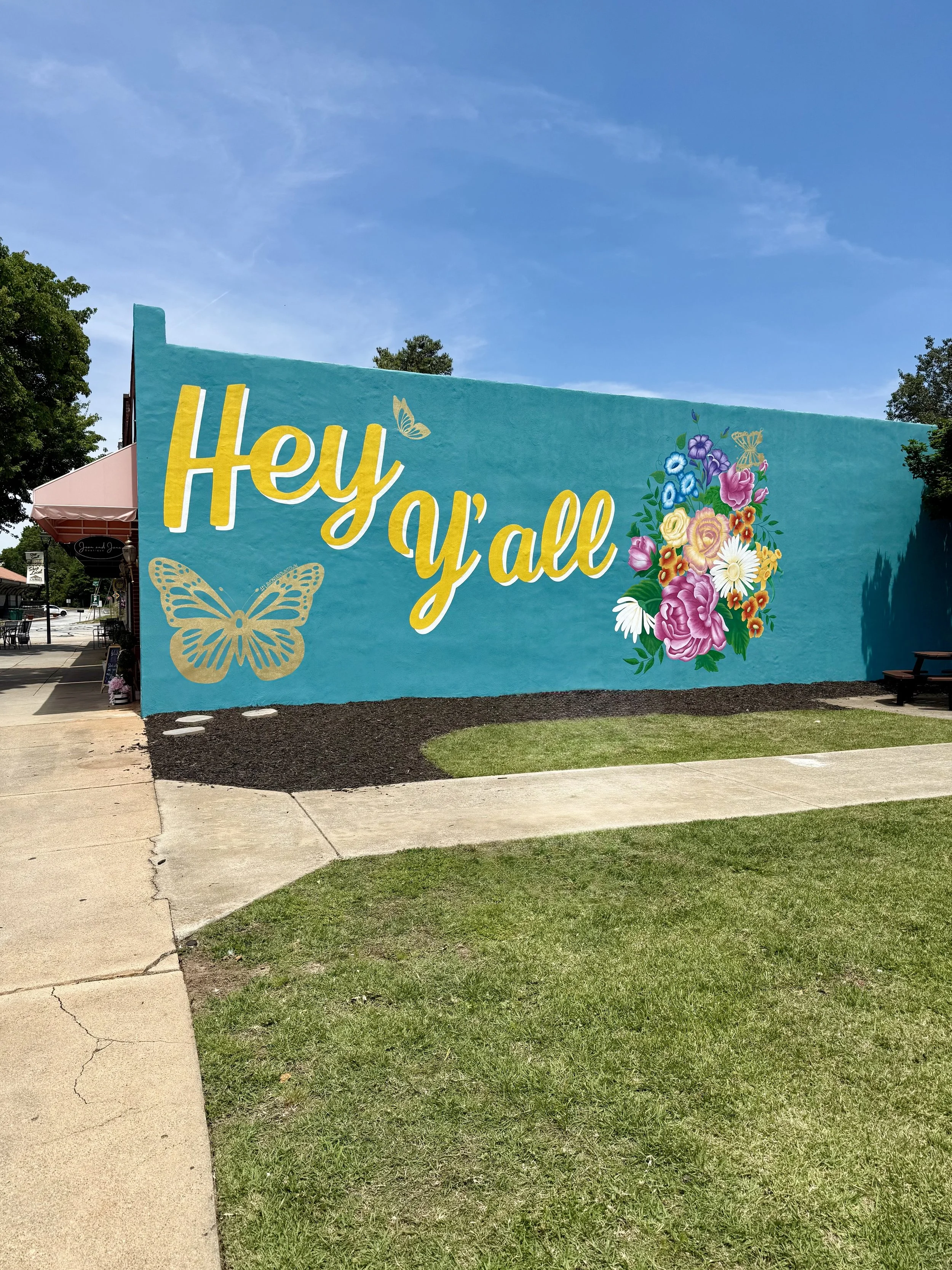 A large outdoor mural with a blue background reading 'Hey y'all' in yellow and white letters, featuring floral and butterfly illustrations.